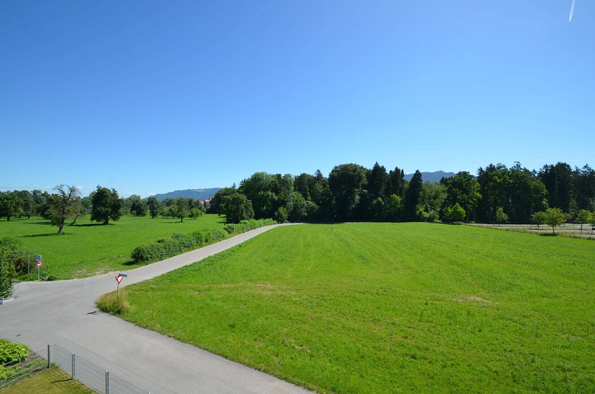 Panoramablick über grüne Felder und eine Landstraße mit bewaldeten Hügeln im Hintergrund.
