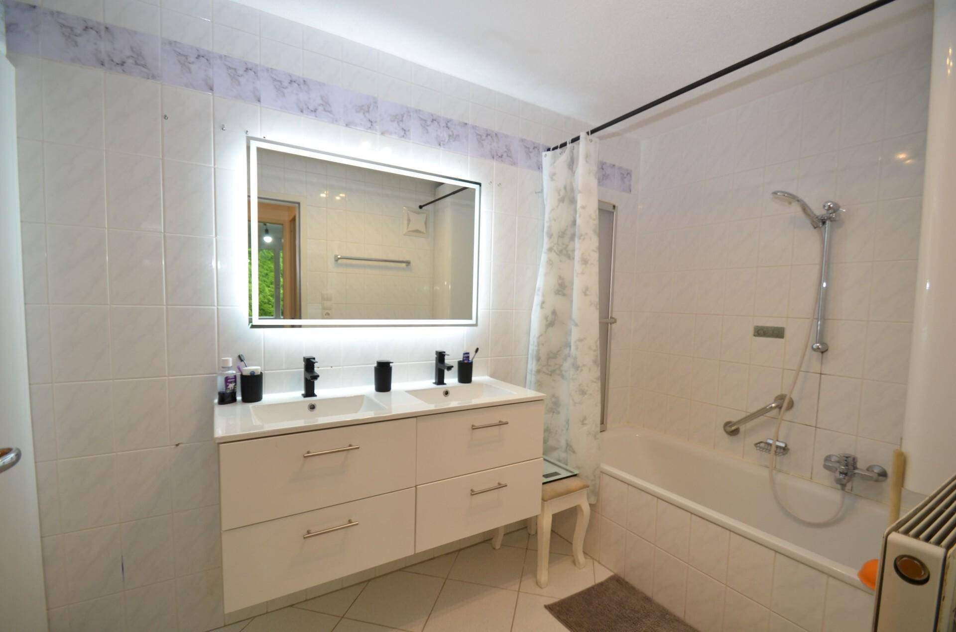 Modernes Badezimmer mit Doppelwaschbecken, beleuchtetem Spiegel und Badewanne mit Duschfunktion.