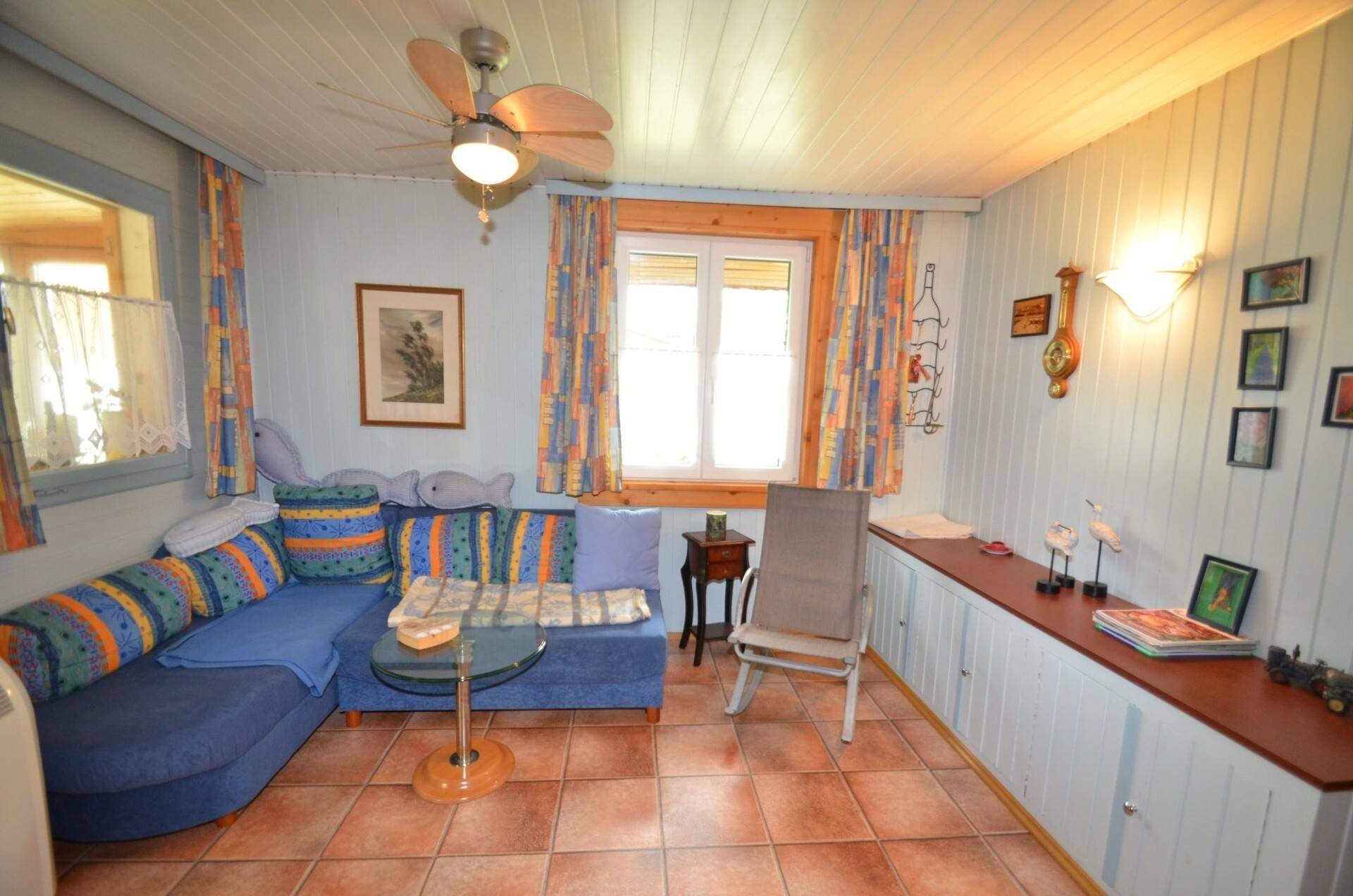 Helles Wohnzimmer mit blauem Ecksofa, Deckenventilator und Terrakottafliesen.