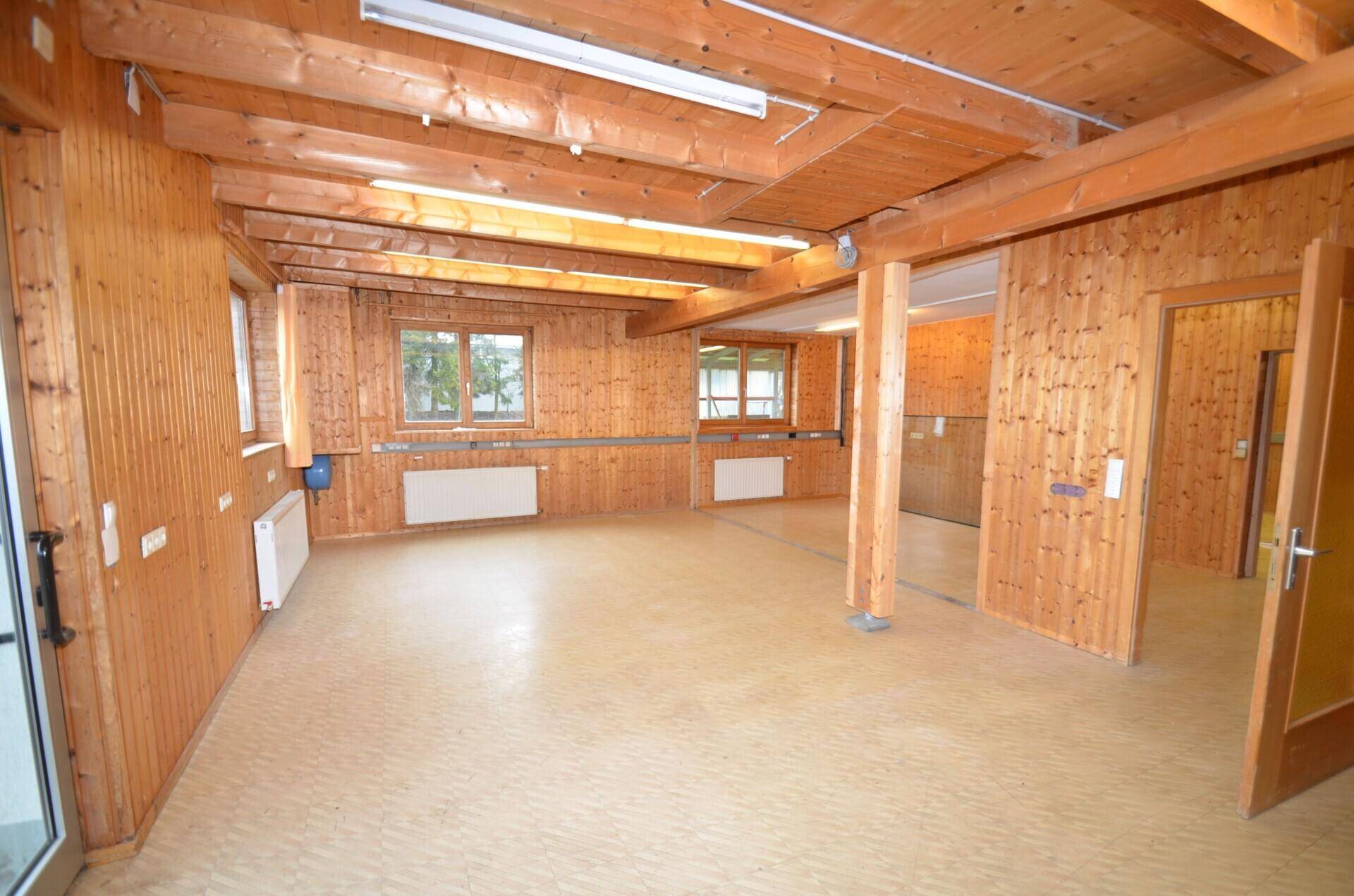 Geräumiger Gewerberaum mit Holzvertäfelung, sichtbaren Deckenbalken und Linoleumboden, ideal für verschiedene Nutzungen.