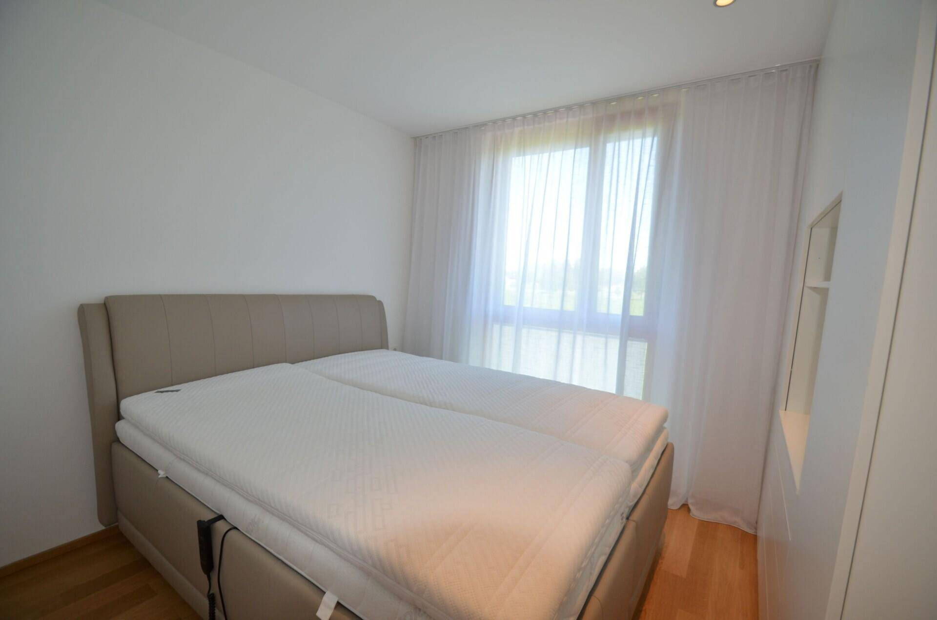 Gemütliches Schlafzimmer mit Doppelbett, Holzboden und hellem Fenster mit Vorhängen.
