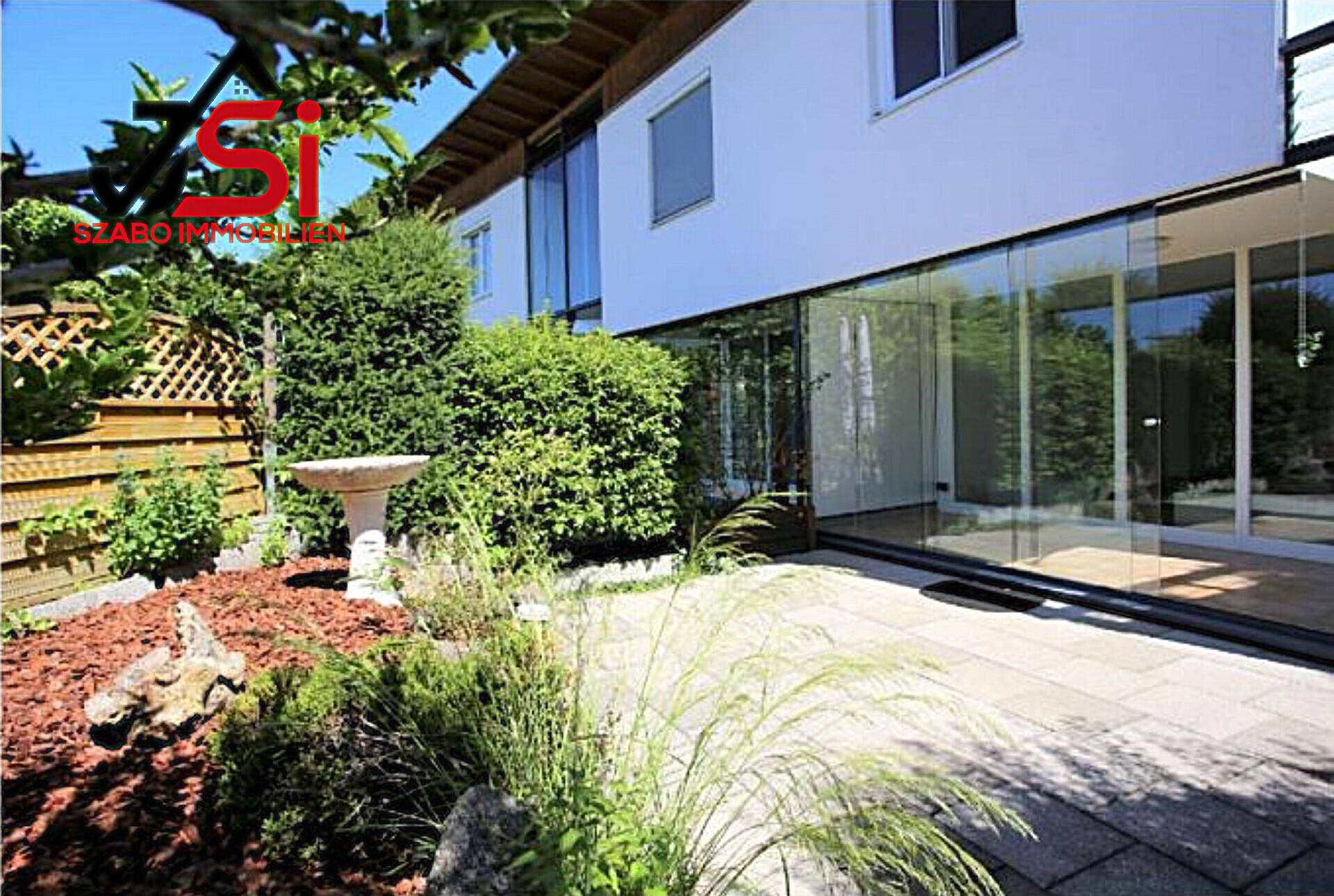 Haus mit moderner Glasfassade, gepflasterter Terrasse und einem gepflegten Gartenbereich mit Vogeltränke.