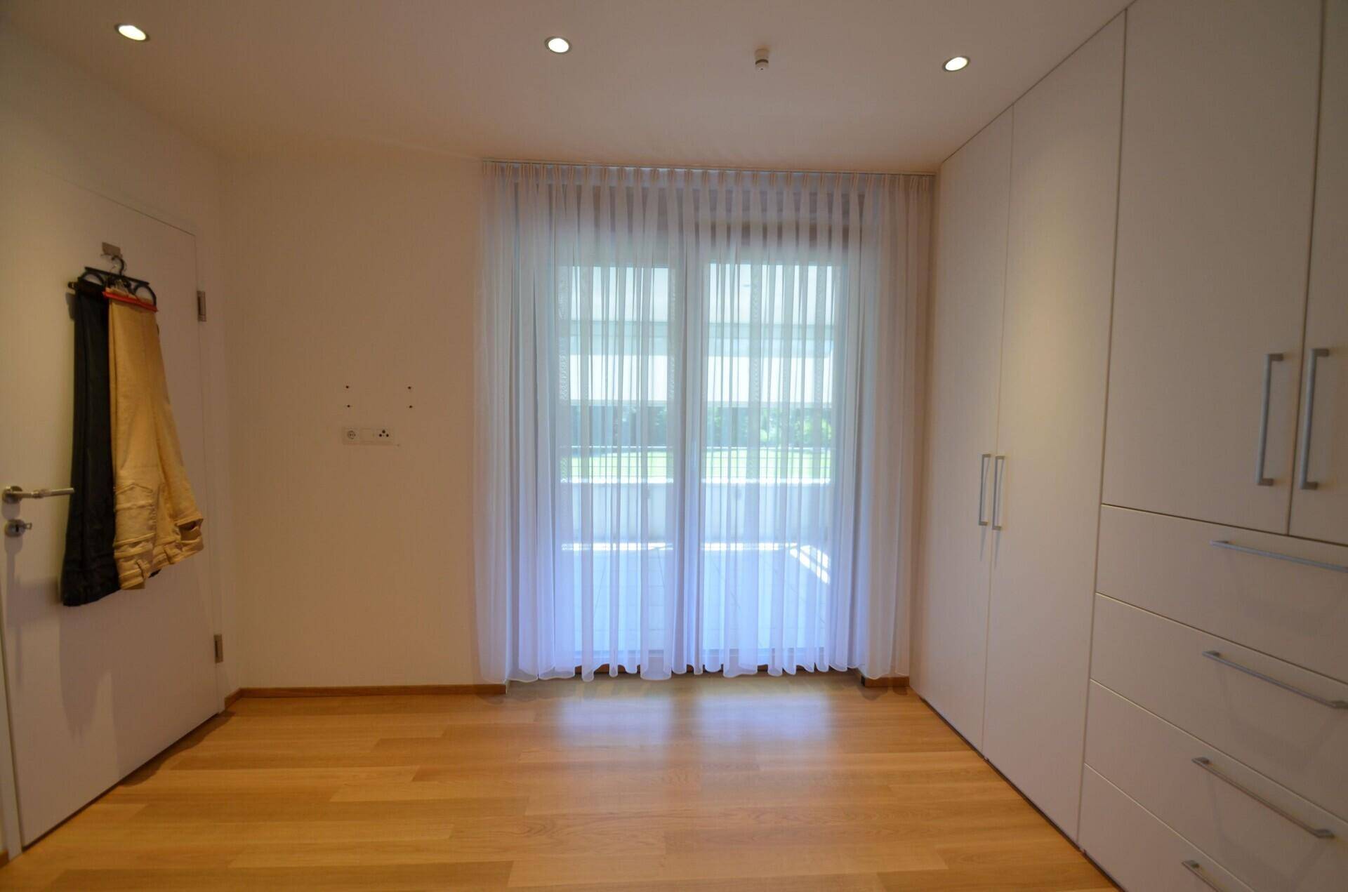 Helles Schlafzimmer mit Holzboden, Einbauschränken und großem Fenster mit Vorhängen.