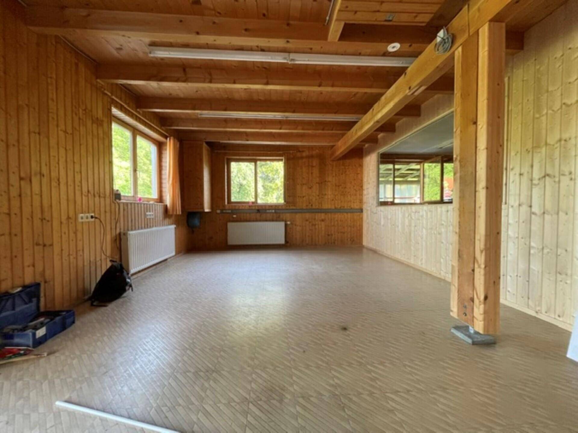 Geräumiger Raum mit Holzvertäfelung, sichtbaren Deckenbalken und mehreren Fenstern für natürliche Belichtung.