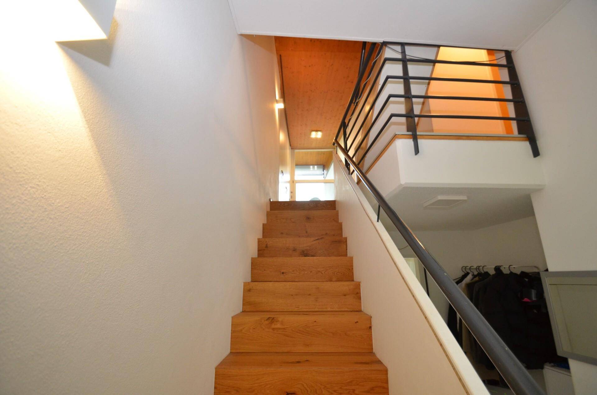 Holztreppe mit Metallgeländer führt zu den oberen Etagen, mit heller Wandgestaltung.