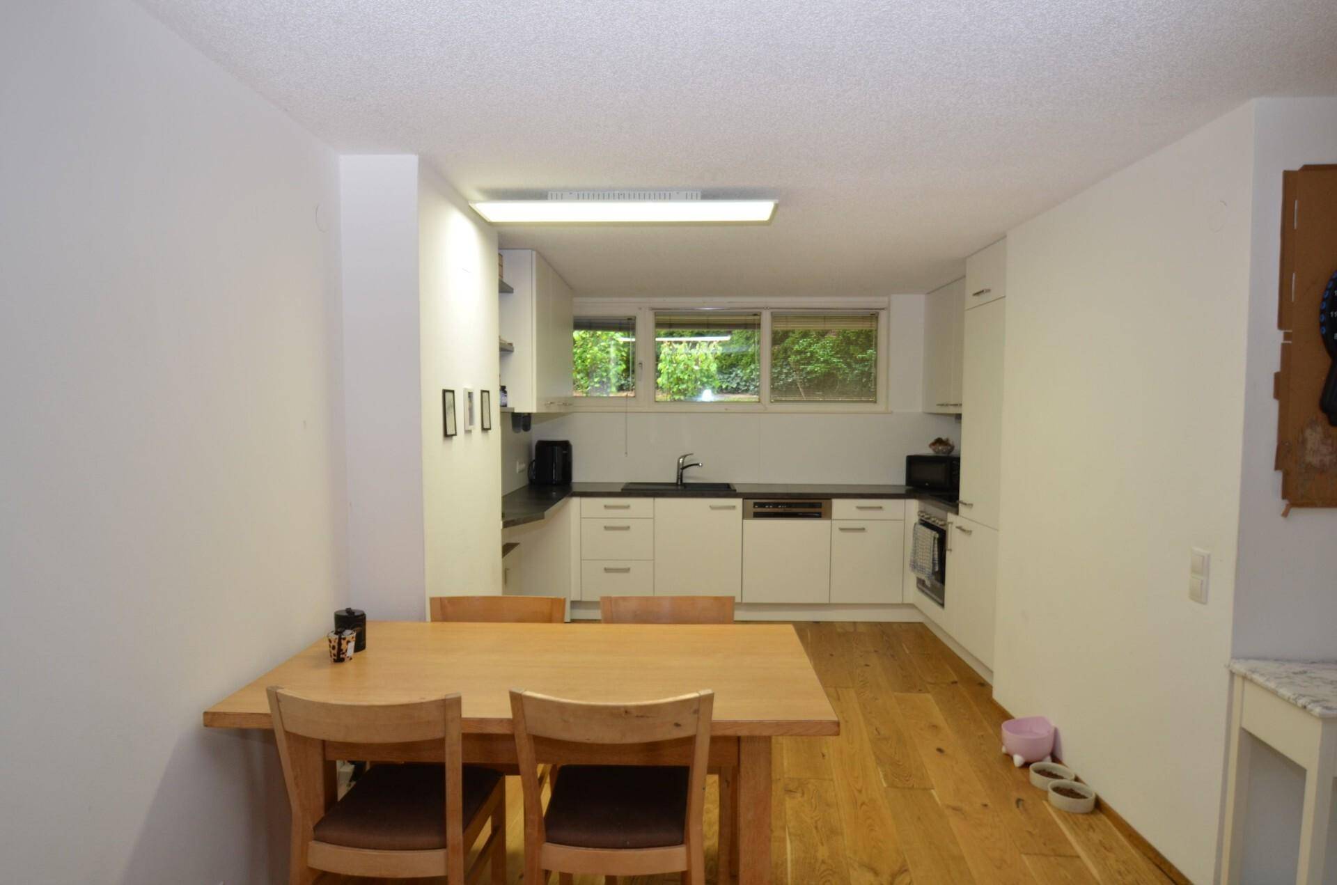 Essbereich mit Holztisch und Stühlen, angrenzend an eine moderne weiße Küche mit Fenster.