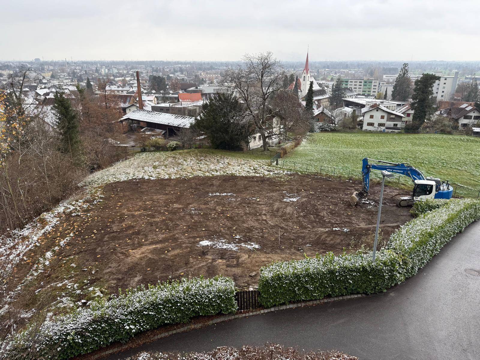 Blick auf ein Baugrundstück mit Bagger und Hecke, umgeben von Häusern und Bäumen.