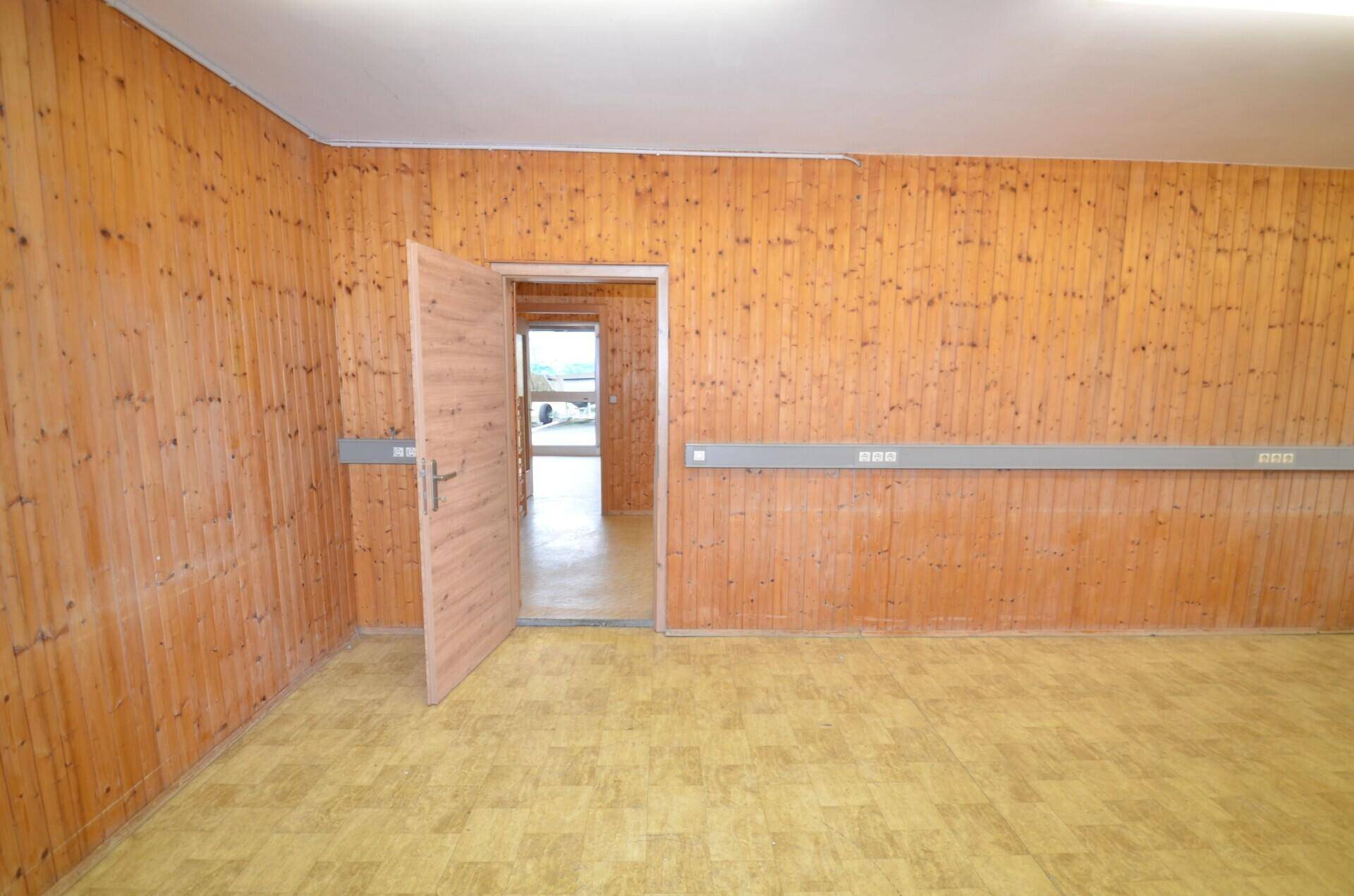 Leerer Raum mit Holzvertäfelung und Linoleumboden, ausgestattet mit Wandsteckdosenleisten und einer Tür zu einem weiteren Bereich.