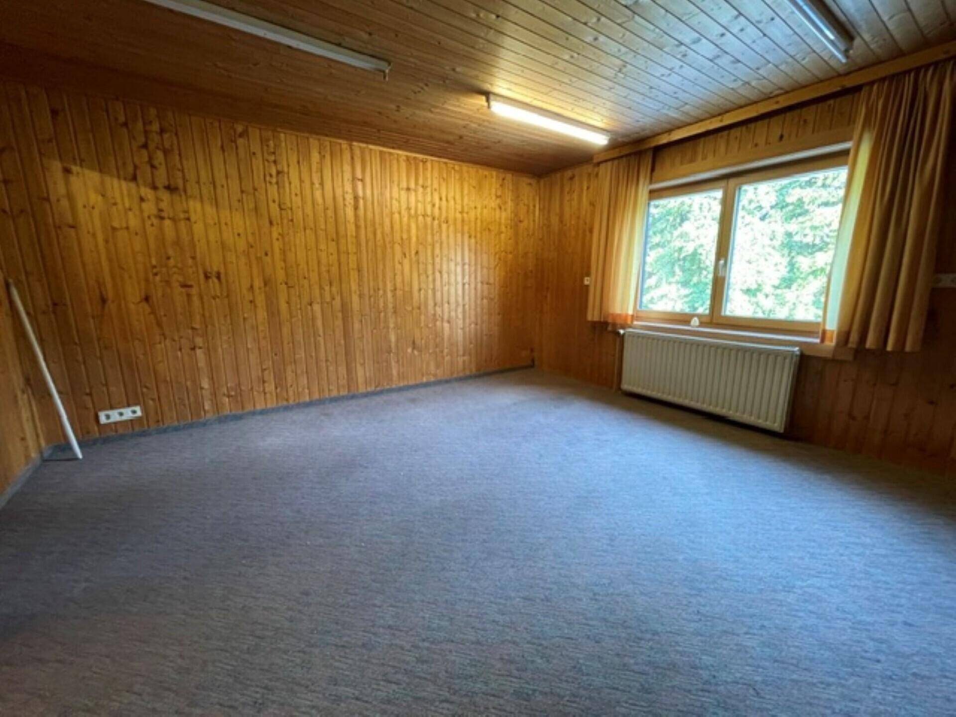Innenraum mit Holzvertäfelung, Fenster mit Vorhängen, Heizkörper und grauem Teppichboden.