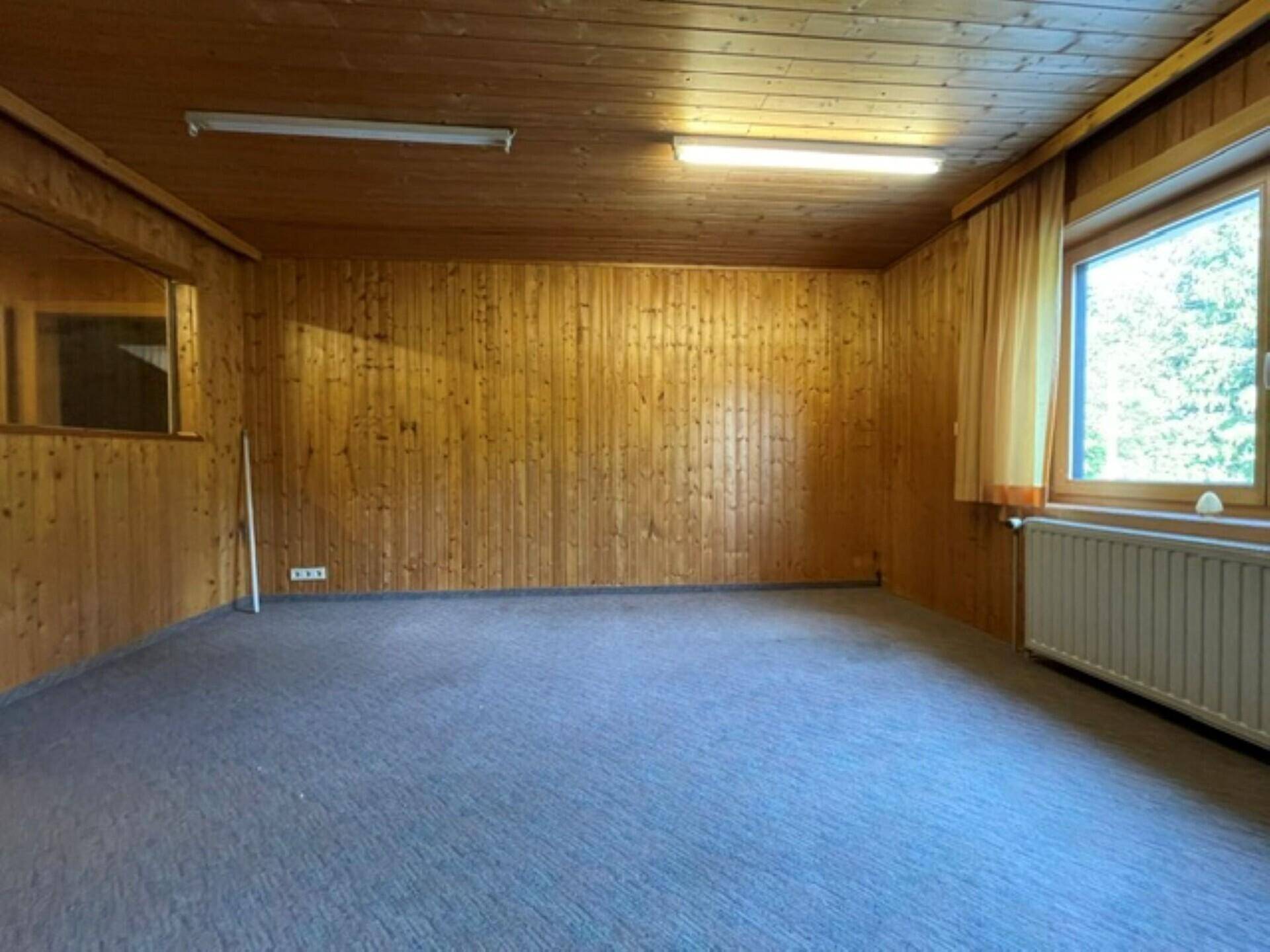 Innenraum mit Holzvertäfelung, grauem Teppichboden, Fenster und einer Spiegelfläche an der Seitenwand.