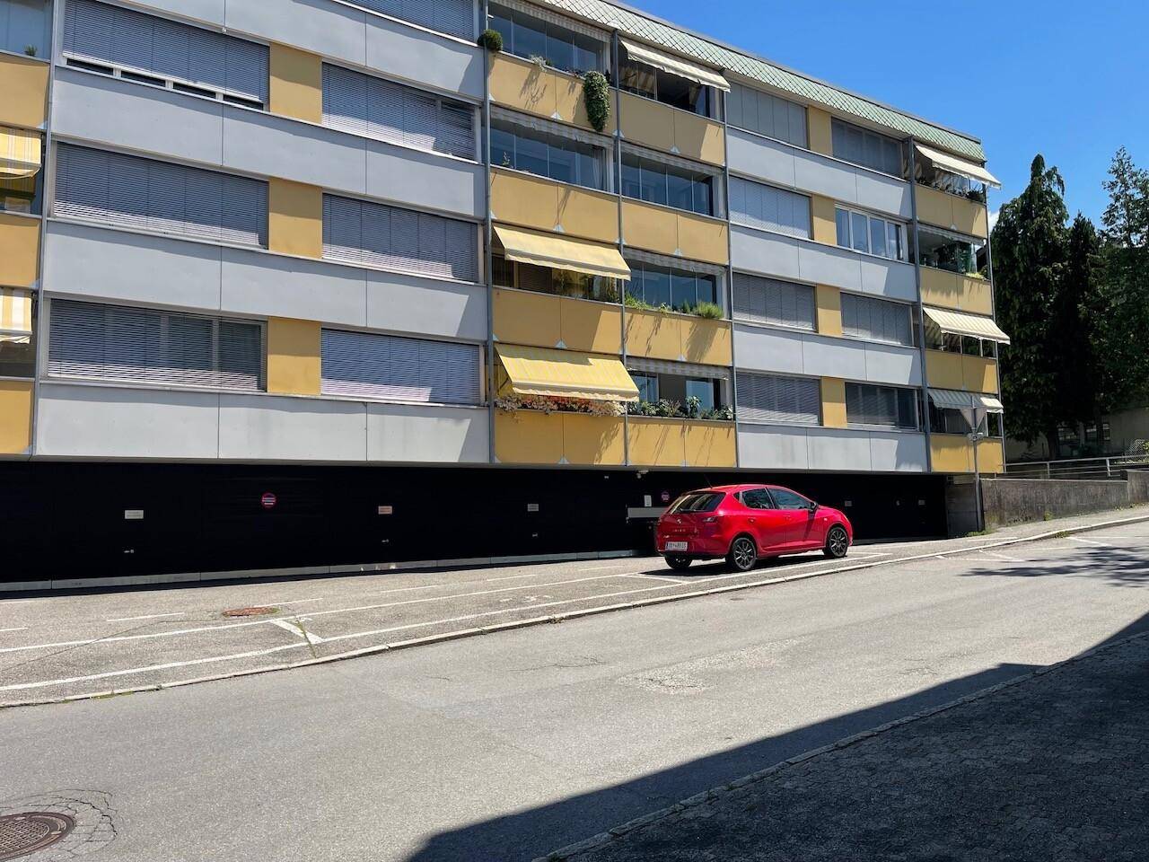 Detailansicht der Gebäudefassade mit gelben Elementen und Markisen, sowie einem geparkten roten Auto davor.
