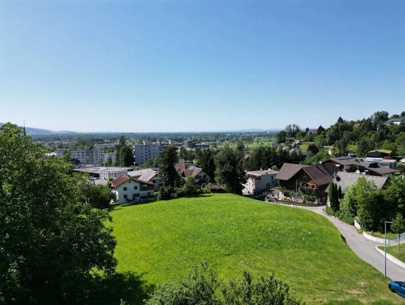 Weitläufige Landschaft mit einer großen grünen Wiese, umgeben von Häusern und Bergen.
