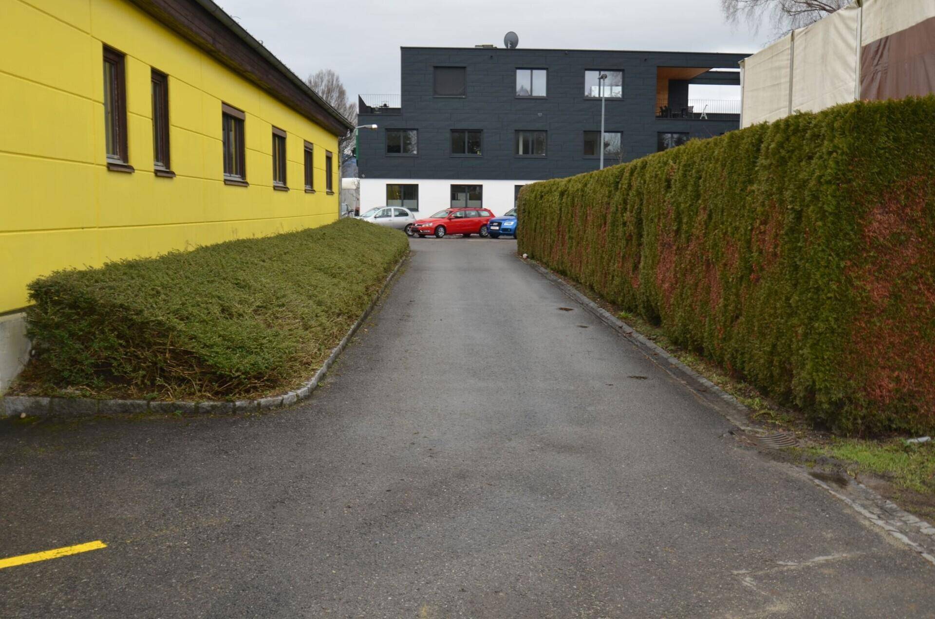 Zufahrtsweg zwischen einem gelben Gebäude und einer hohen Hecke, mit einem modernen Gebäude im Hintergrund.