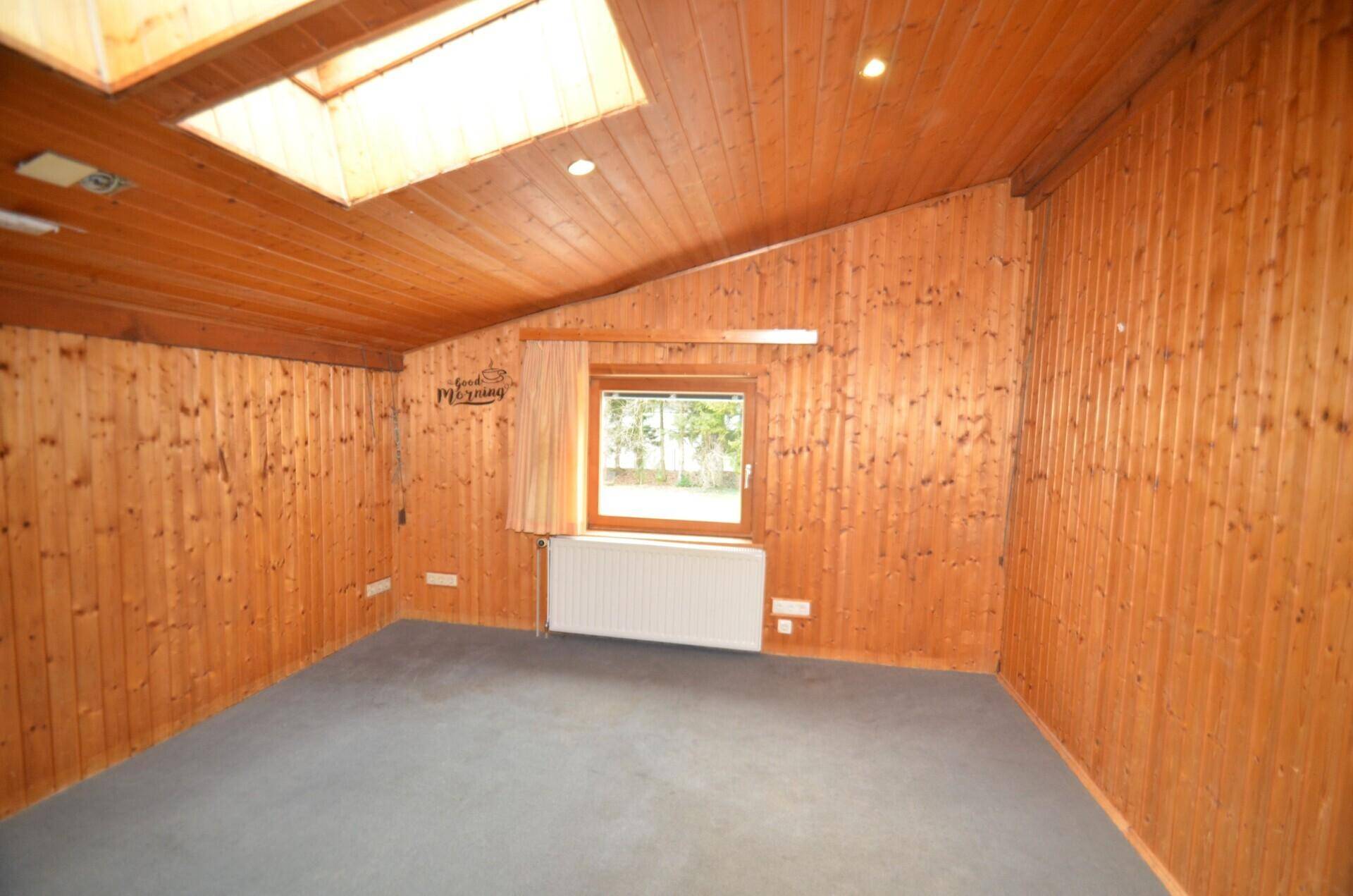 Gemütlicher Raum mit Holzvertäfelung, Teppichboden, Dachfenster und einem weiteren Fenster mit Blick ins Grüne.