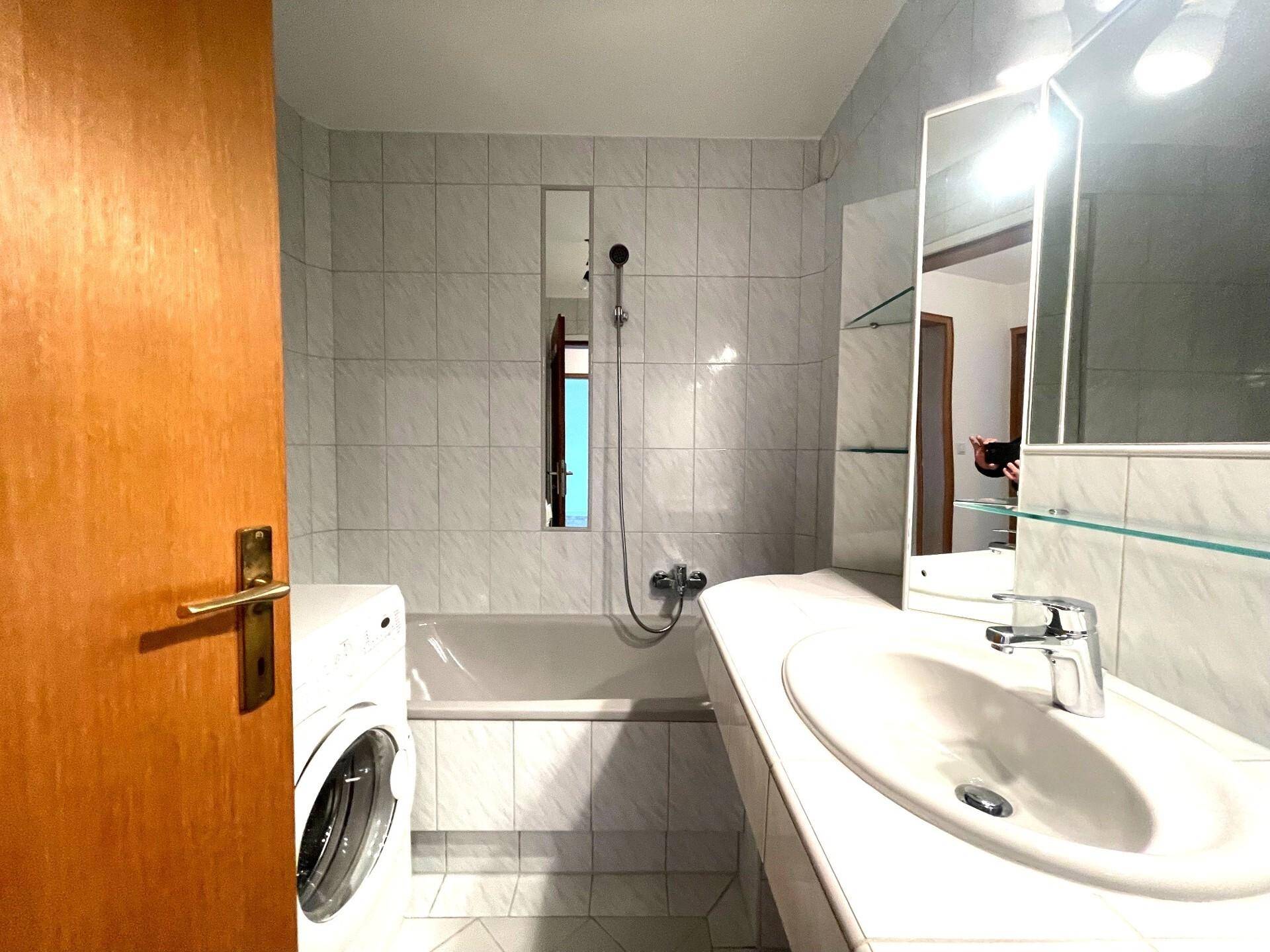Gefliestes Badezimmer mit Badewanne, Waschbecken und Spiegel, inklusive Waschmaschine.