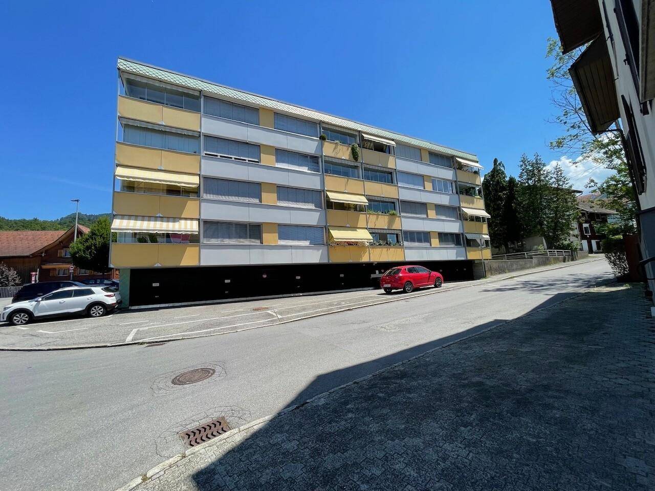 Mehrstöckiges Wohngebäude mit gelben Markisen und Parkplätzen an der Straße, eingebettet in eine grüne Umgebung.