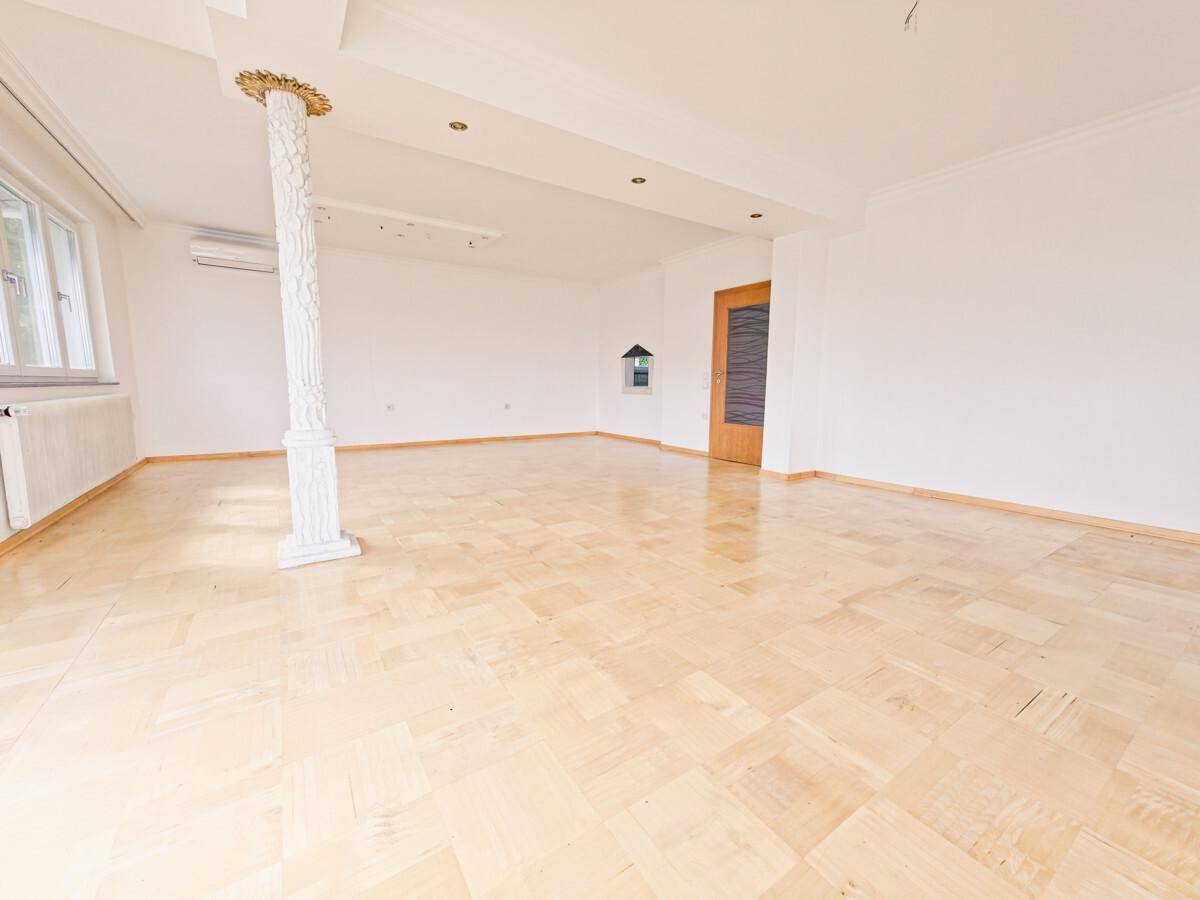 Großes, leeres Wohnzimmer mit Parkettboden, dekorativer Säule und viel Tageslicht.