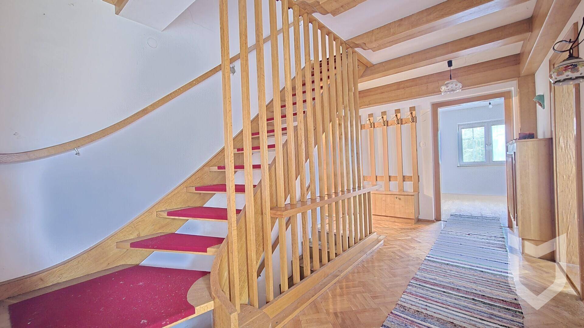 Heller Flur mit einer geschwungenen Holztreppe, rotem Teppich und sichtbaren Holzbalken an der Decke.