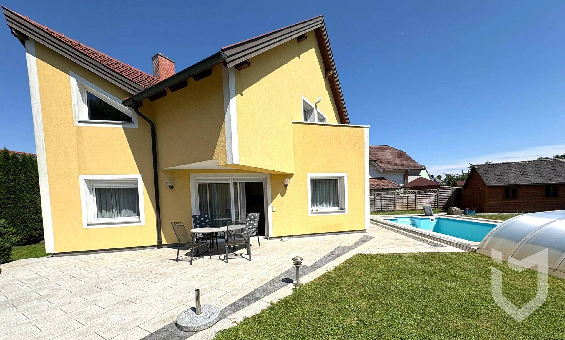 Gelbes Einfamilienhaus mit Terrasse, Pool und Gartenbereich unter strahlend blauem Himmel.