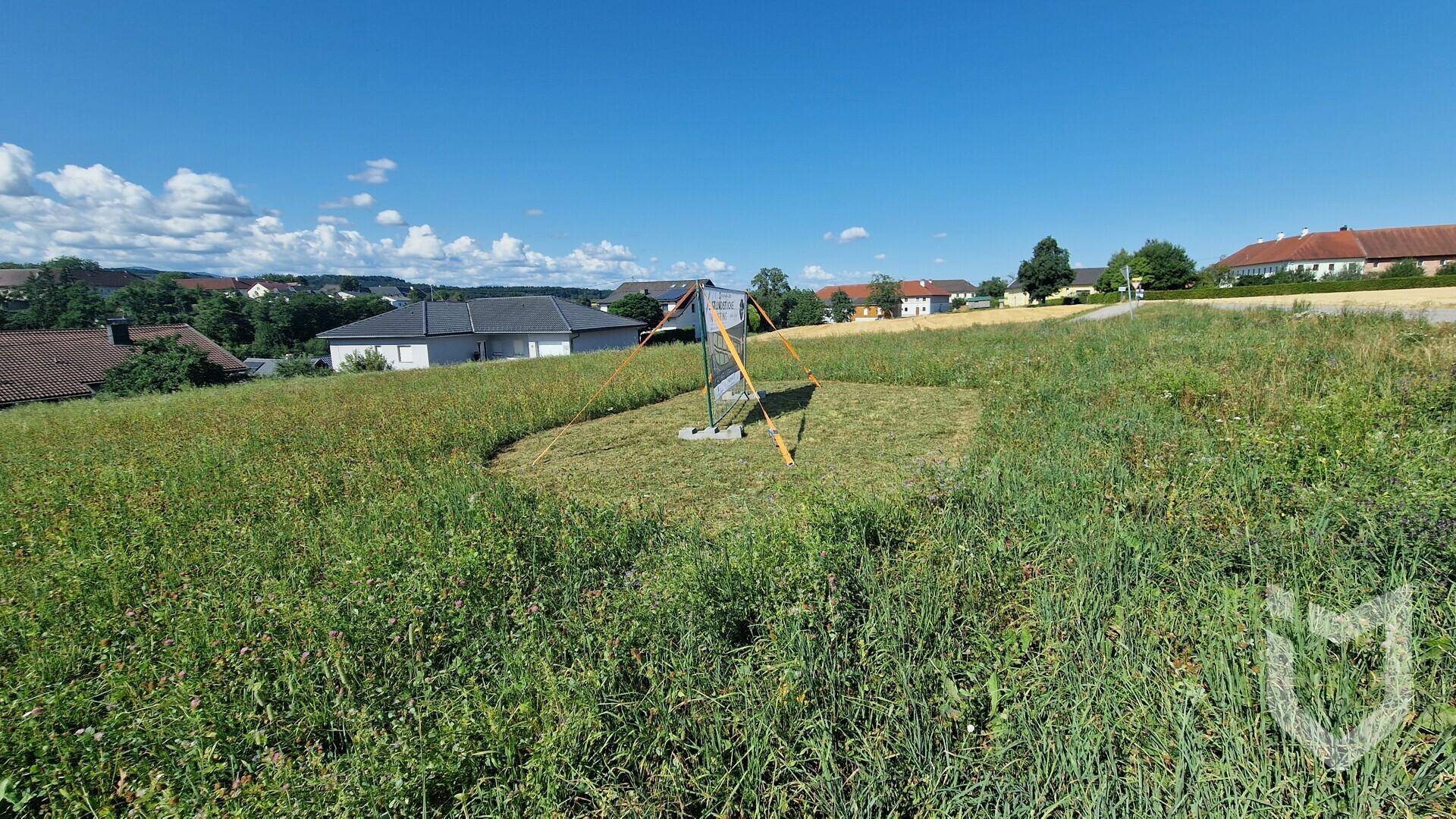 Grüne Wiesenfläche mit einem kleinen Markierungspfahl und Häusern in der Ferne unter strahlend blauem Himmel.