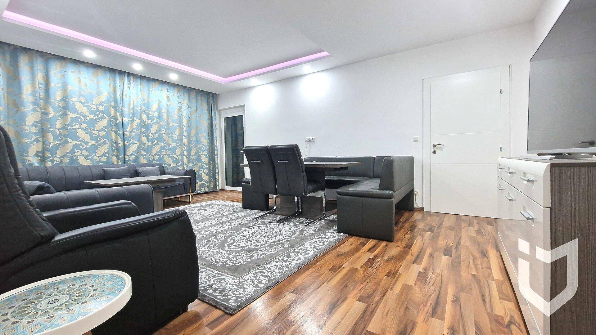 Wohn- und Essbereich mit dunklem Ecksofa, Esstisch und gemusterten Vorhängen.
