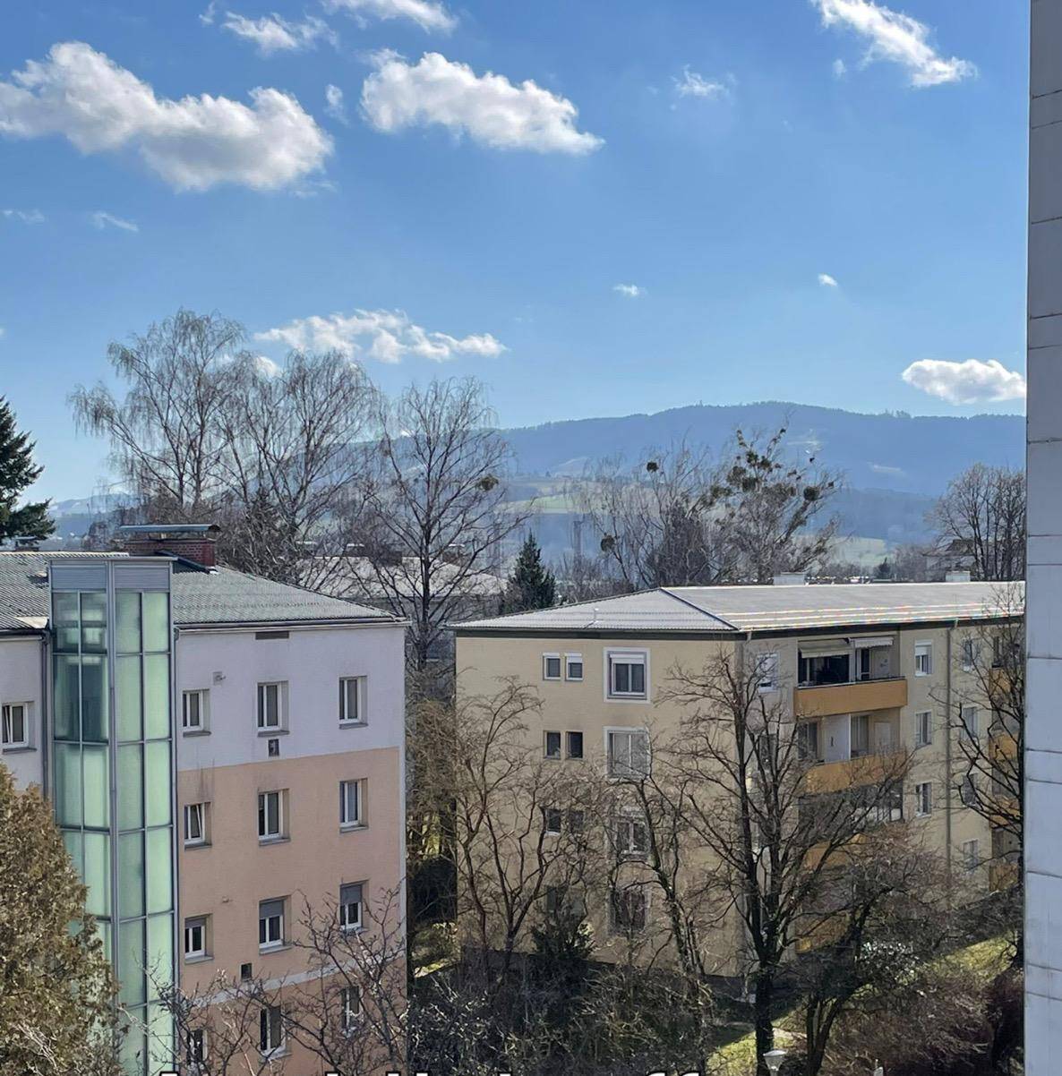 Blick aus dem Fenster auf Wohngebäude, Bäume und ferne Berge unter blauem Himmel.