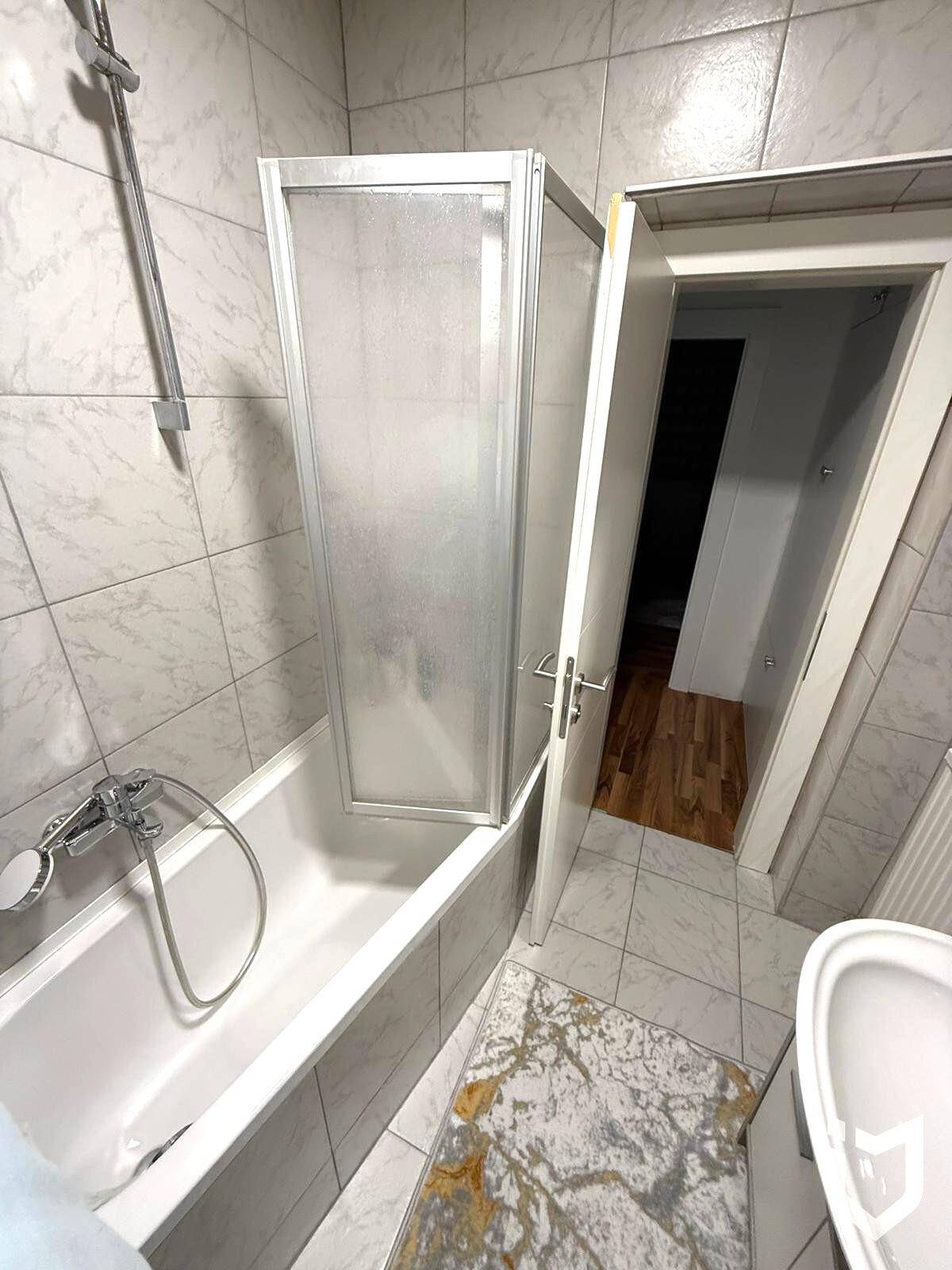 Badezimmer mit Badewanne, Duschabtrennung und WC, komplett gefliest.