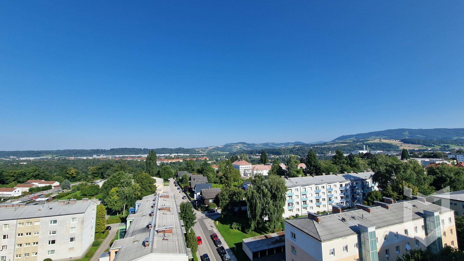 Weitläufiger Blick über eine Wohngegend mit Gebäuden, Bäumen und fernen Hügeln.