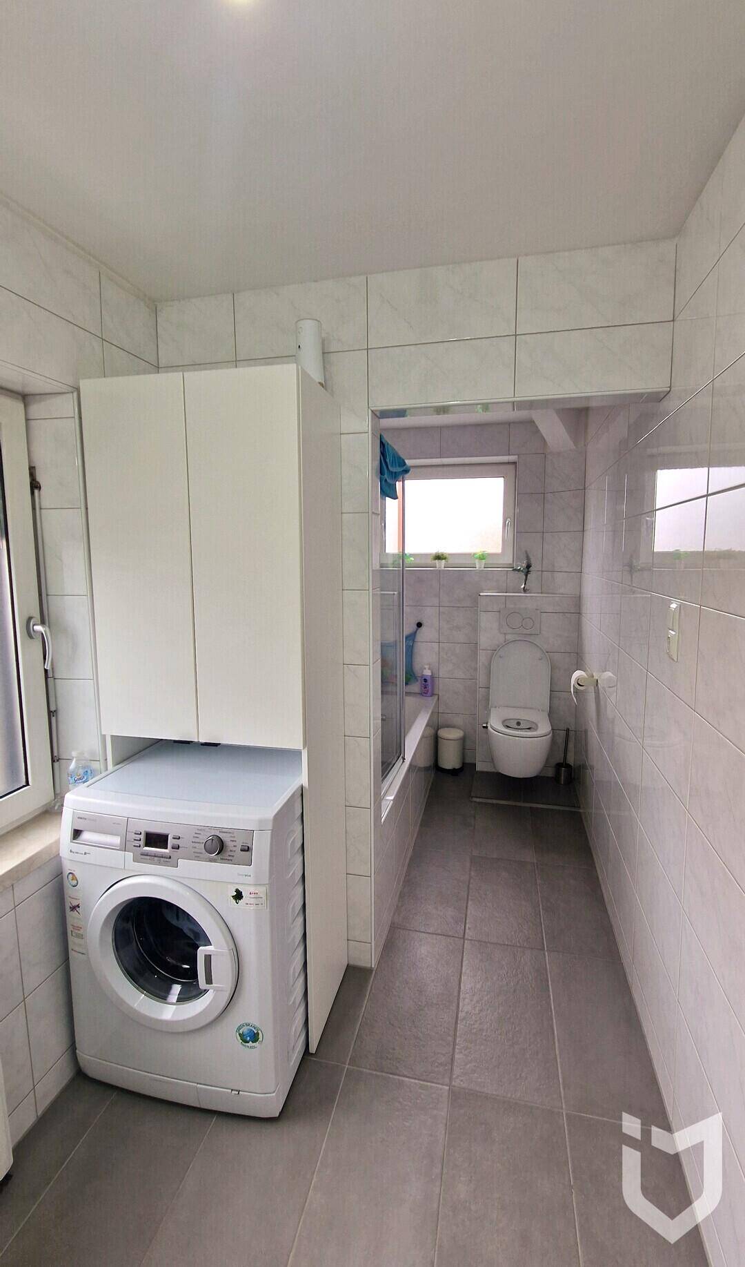 Funktionales Badezimmer mit Waschmaschine, Toilette und Duschbereich, hell gefliest.