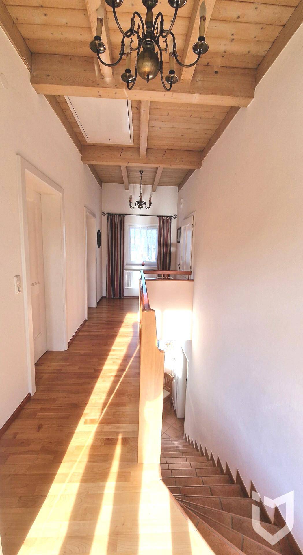 Heller Flur mit Holztreppe, Holzbalkendecke und Kronleuchter, durchflutet von Sonnenlicht.