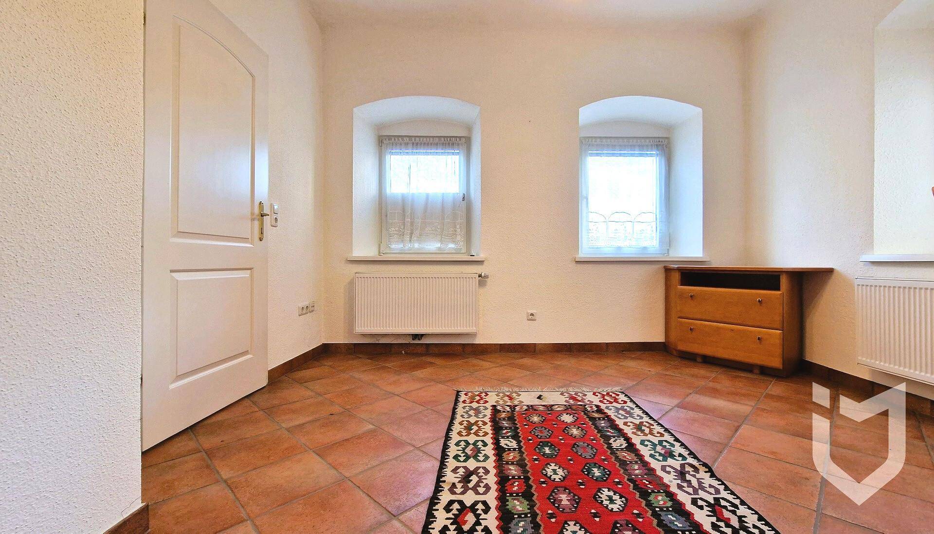 Heller Raum mit Terrakotta-Fliesenboden, zwei Fenstern und einem kleinen Holzschrank.
