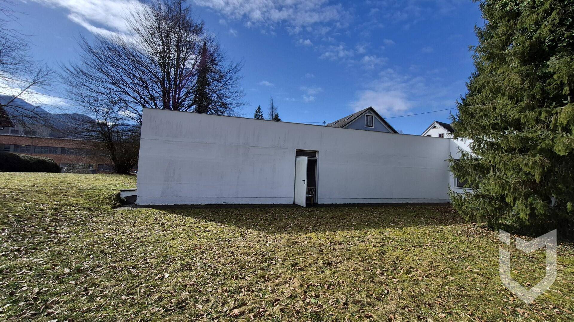 Minimalistisches weißes Gebäude mit einer Tür, umgeben von Rasen und Bäumen vor Bergkulisse.