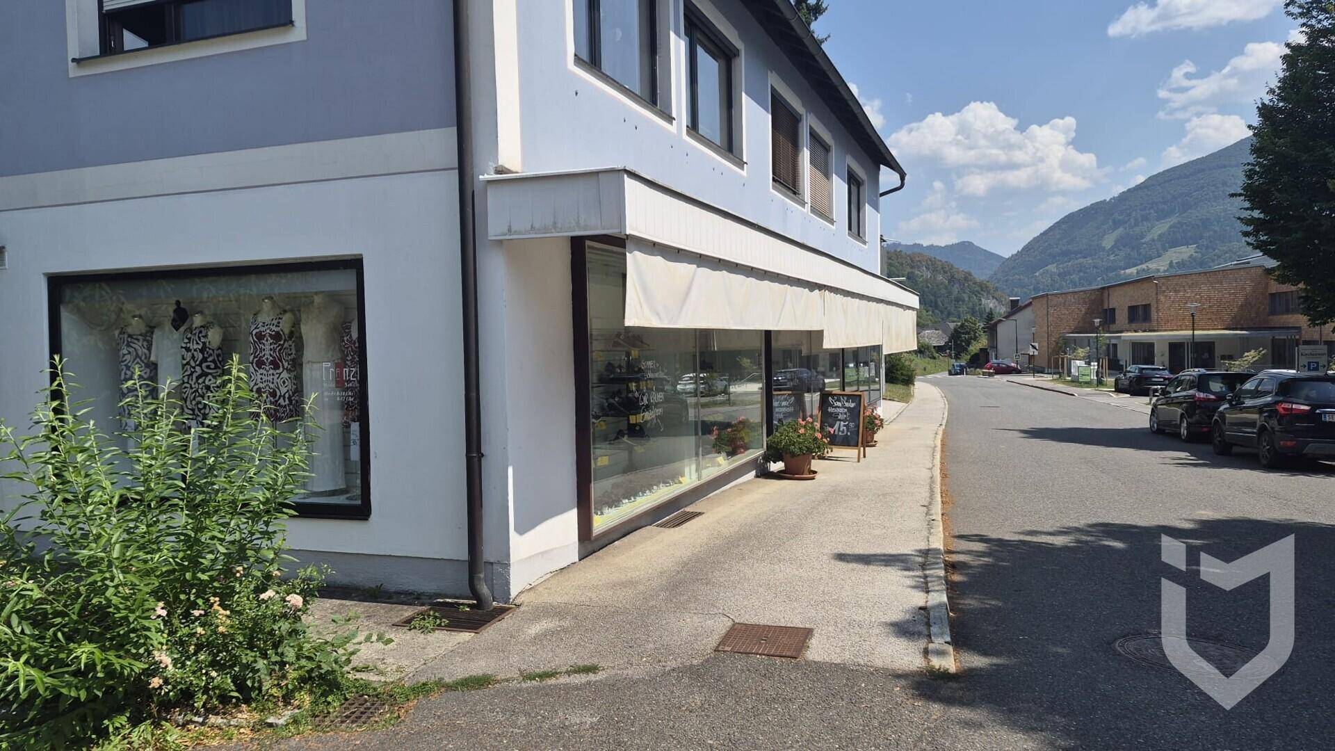 Ansicht eines Gewerbegebäudes mit Schaufenstern an einer Straße, umgeben von Bergen.