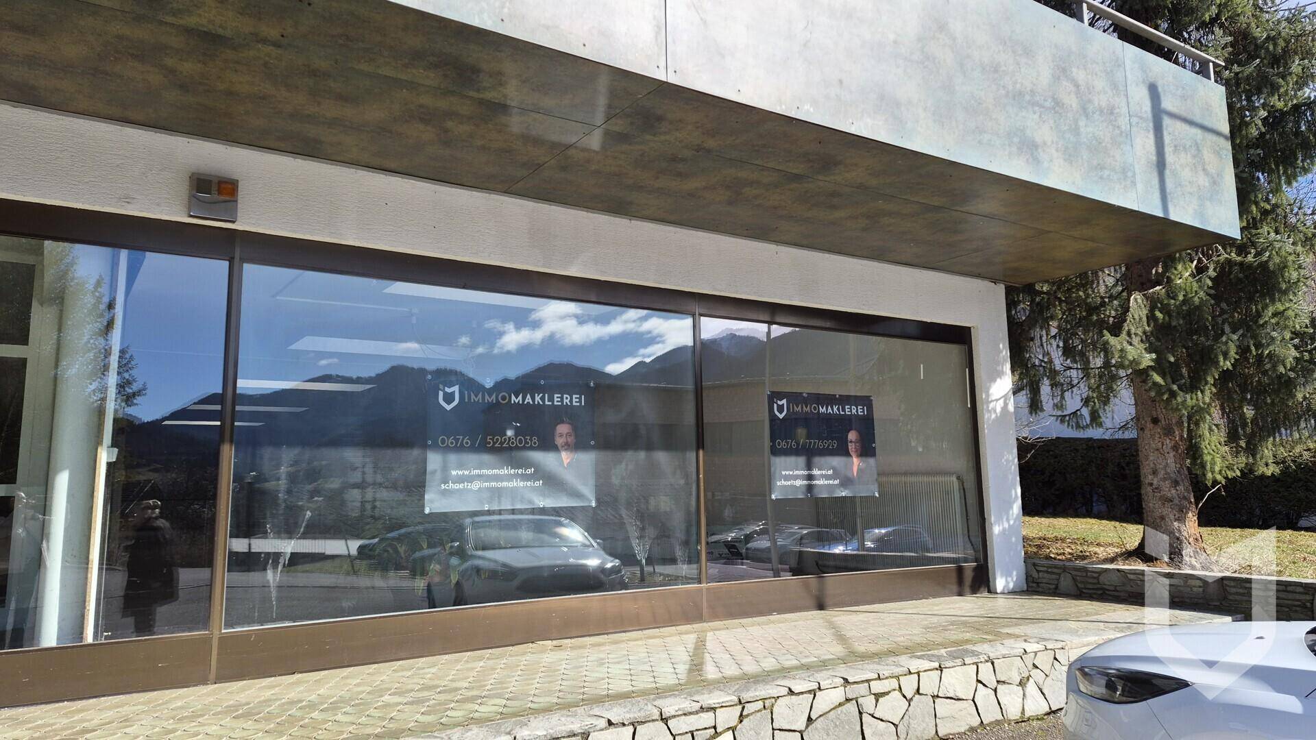 Nahaufnahme der großen Glasfenster einer Gewerbeimmobilie mit Bergreflexionen und Maklerwerbung.