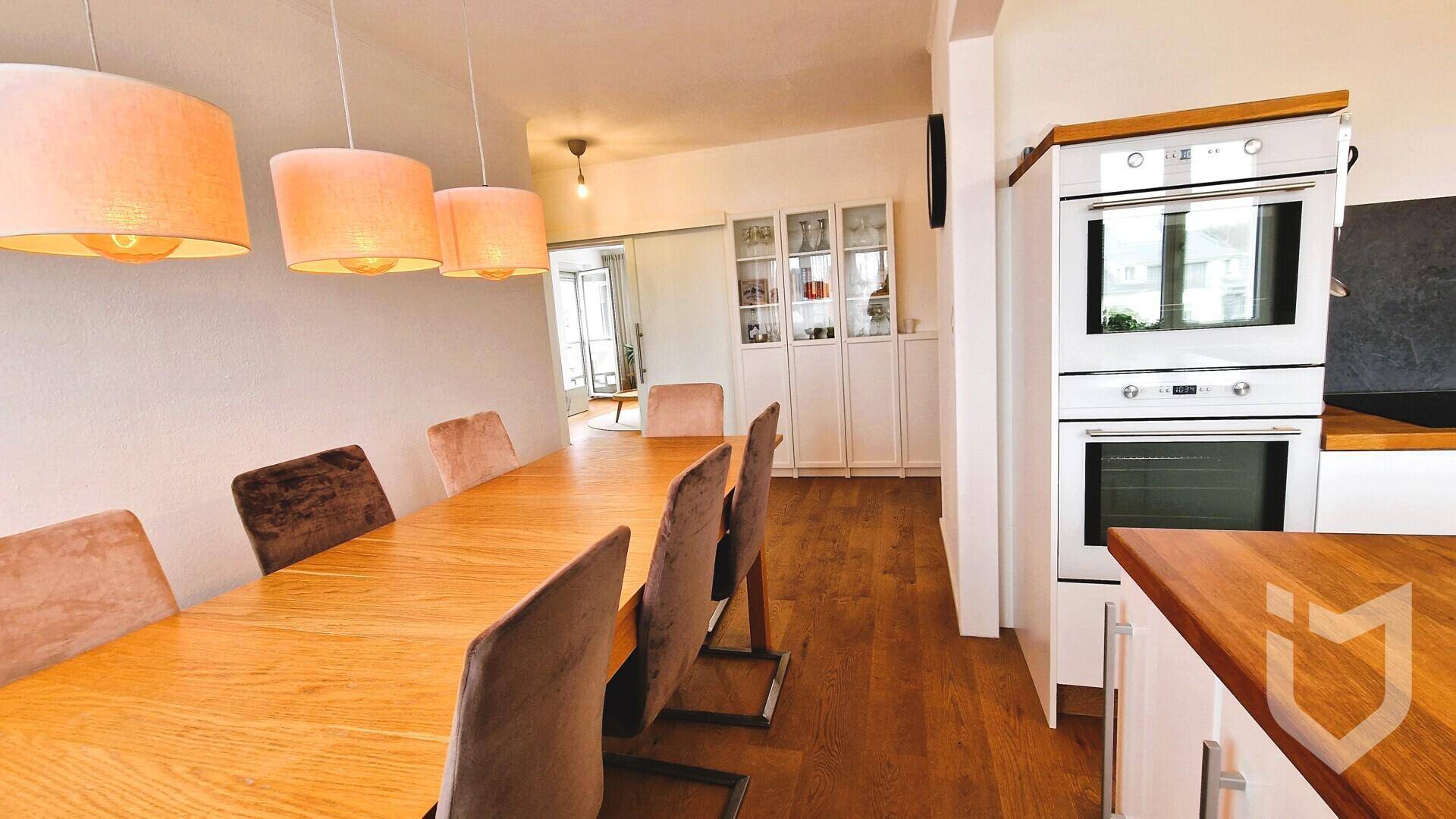 Essbereich mit Holztisch und Blick in die moderne Küche mit Einbaugeräten.