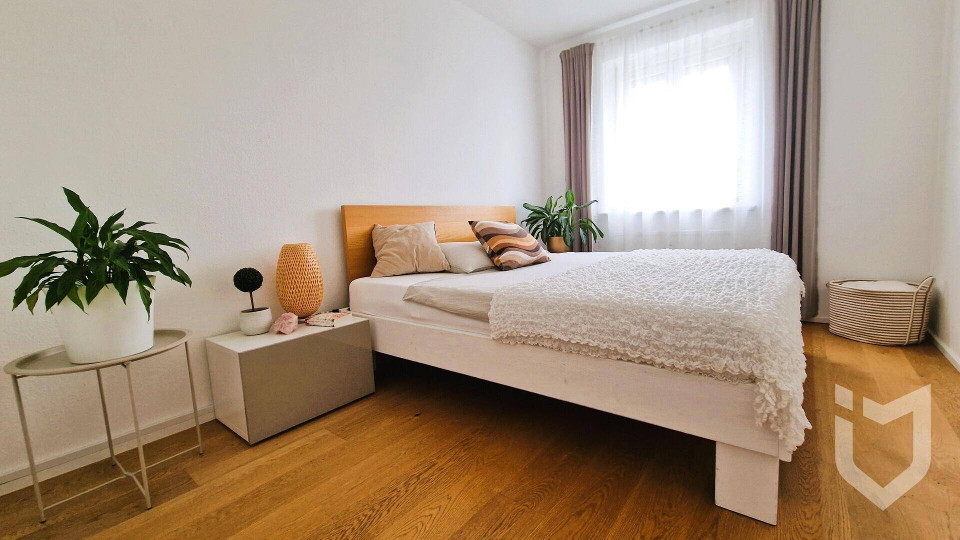 Helles Schlafzimmer mit Doppelbett, Holzboden und dekorativen Pflanzen am Fenster.