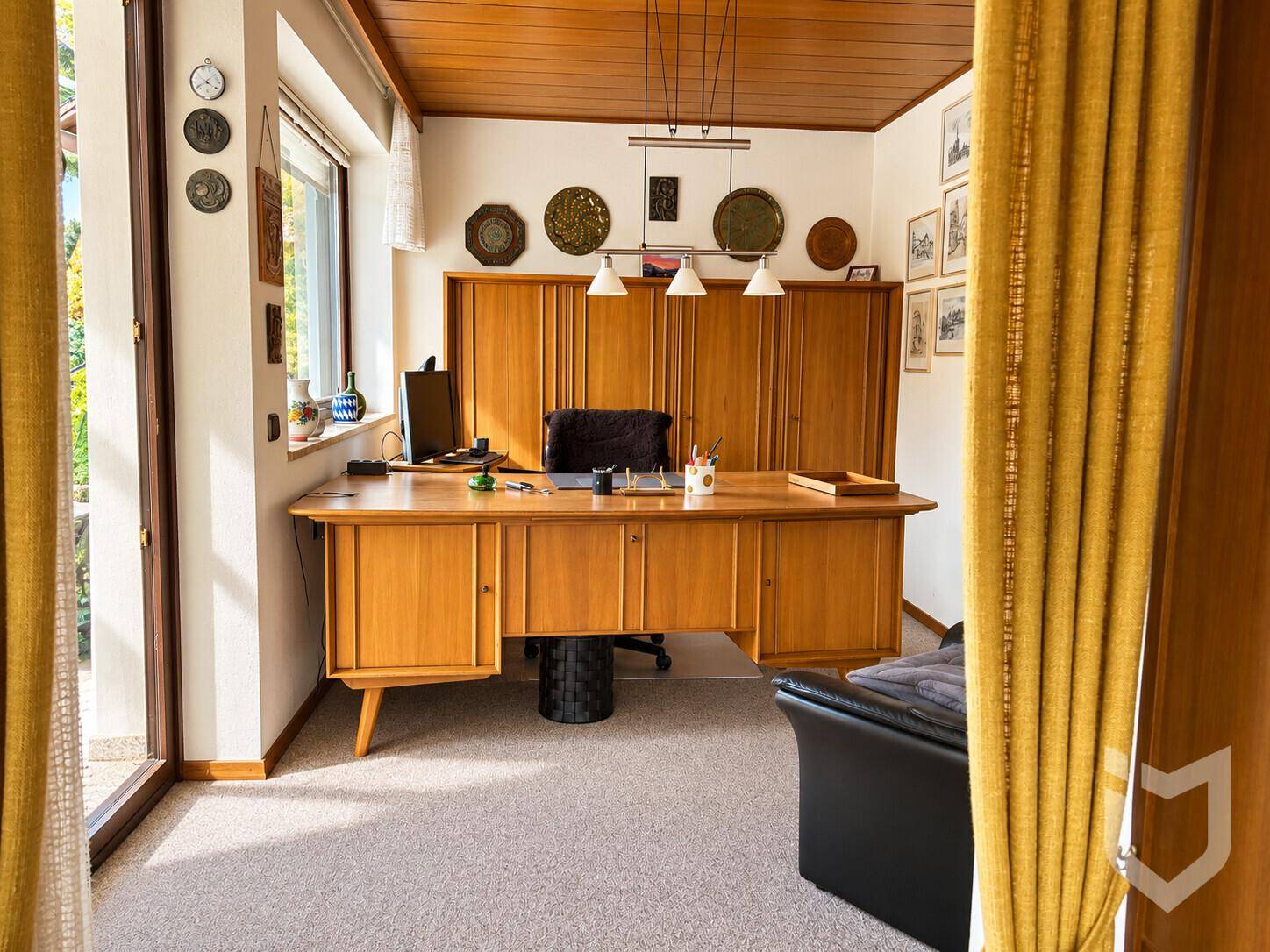 Helles Arbeitszimmer mit Teppichboden, Holzdecke und großem Schreibtisch mit Blick ins Grüne.