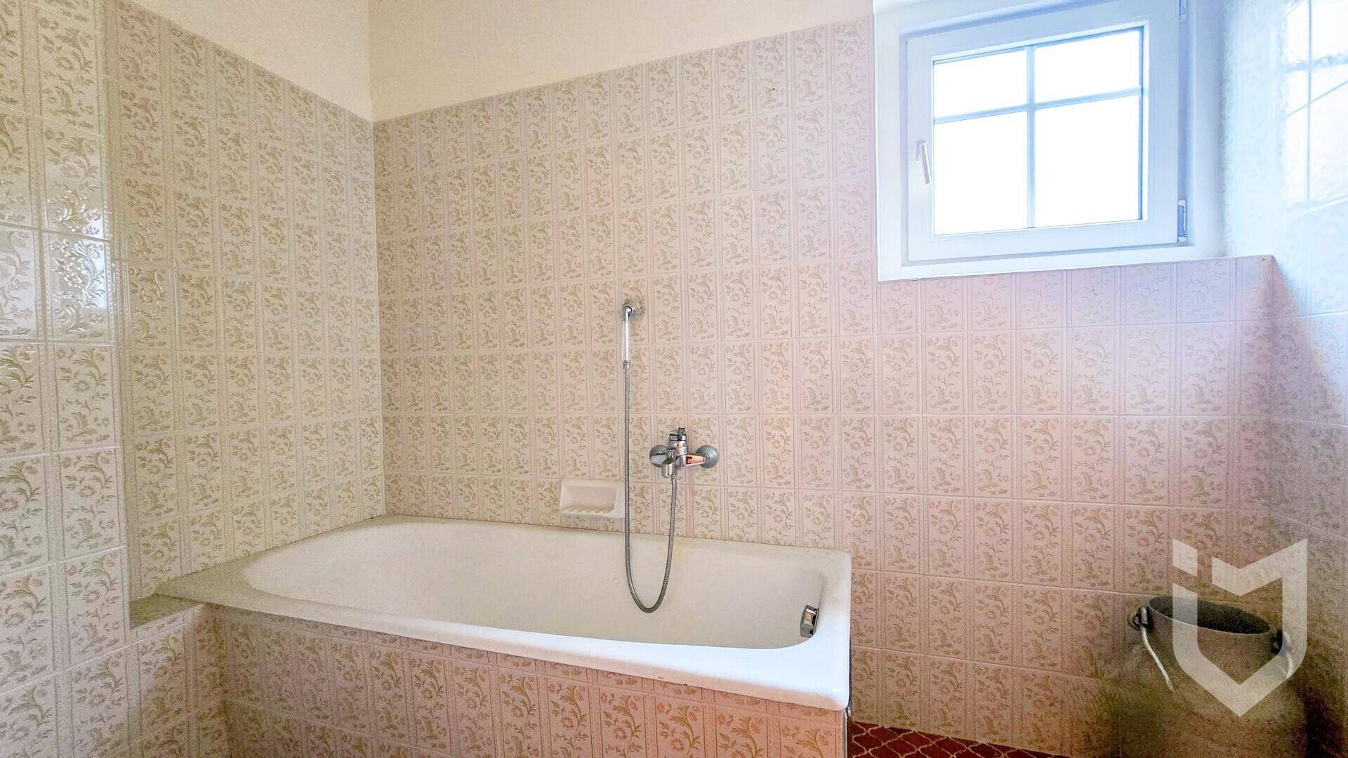 Badezimmer mit eingebauter Badewanne, gemusterten Fliesen und einem Fenster, das eine Renovierung benötigt.