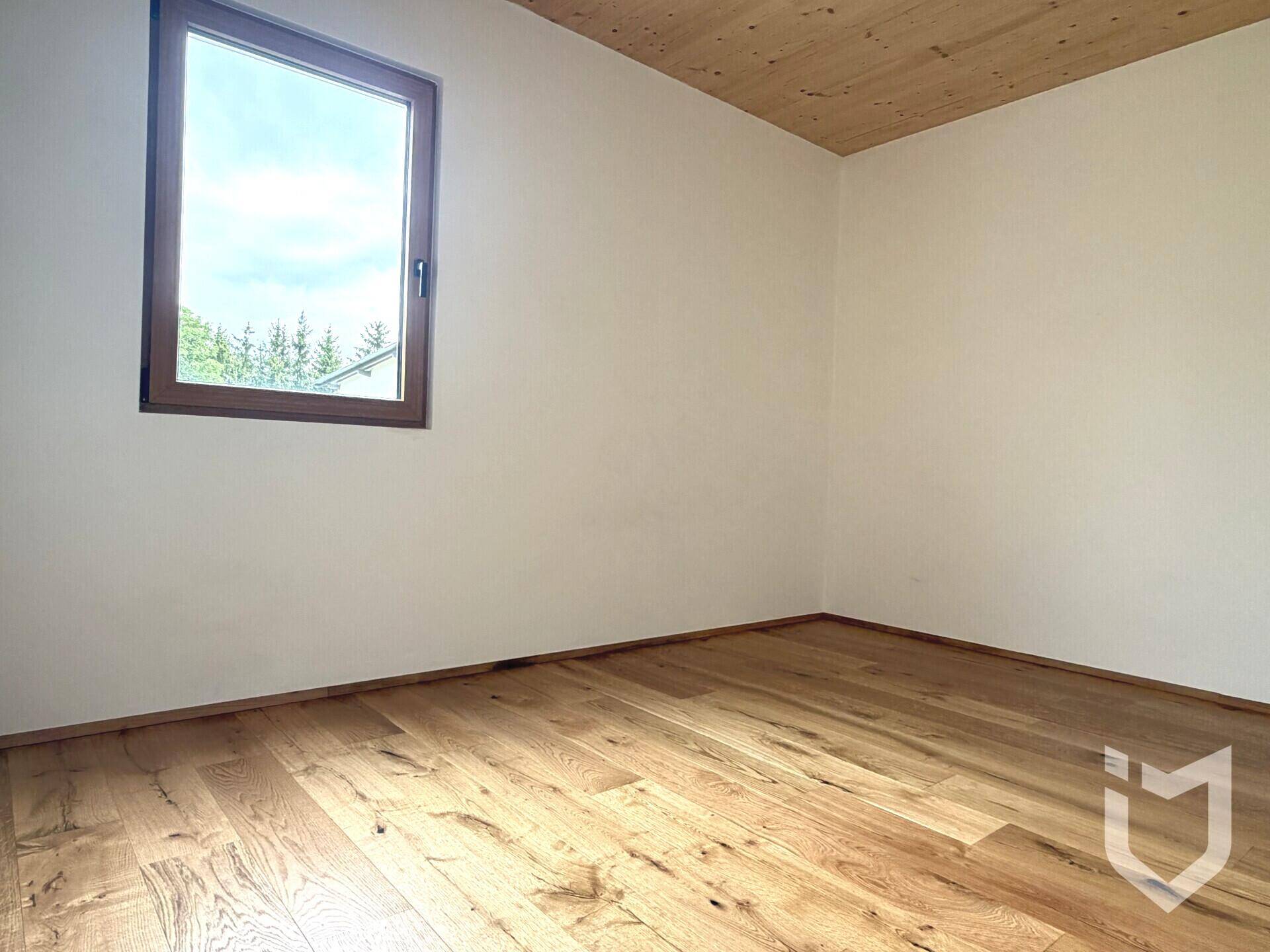 Gemütliches Zimmer mit Holzboden und Holzdecke, einem Fenster mit Blick auf Bäume und viel Tageslicht.