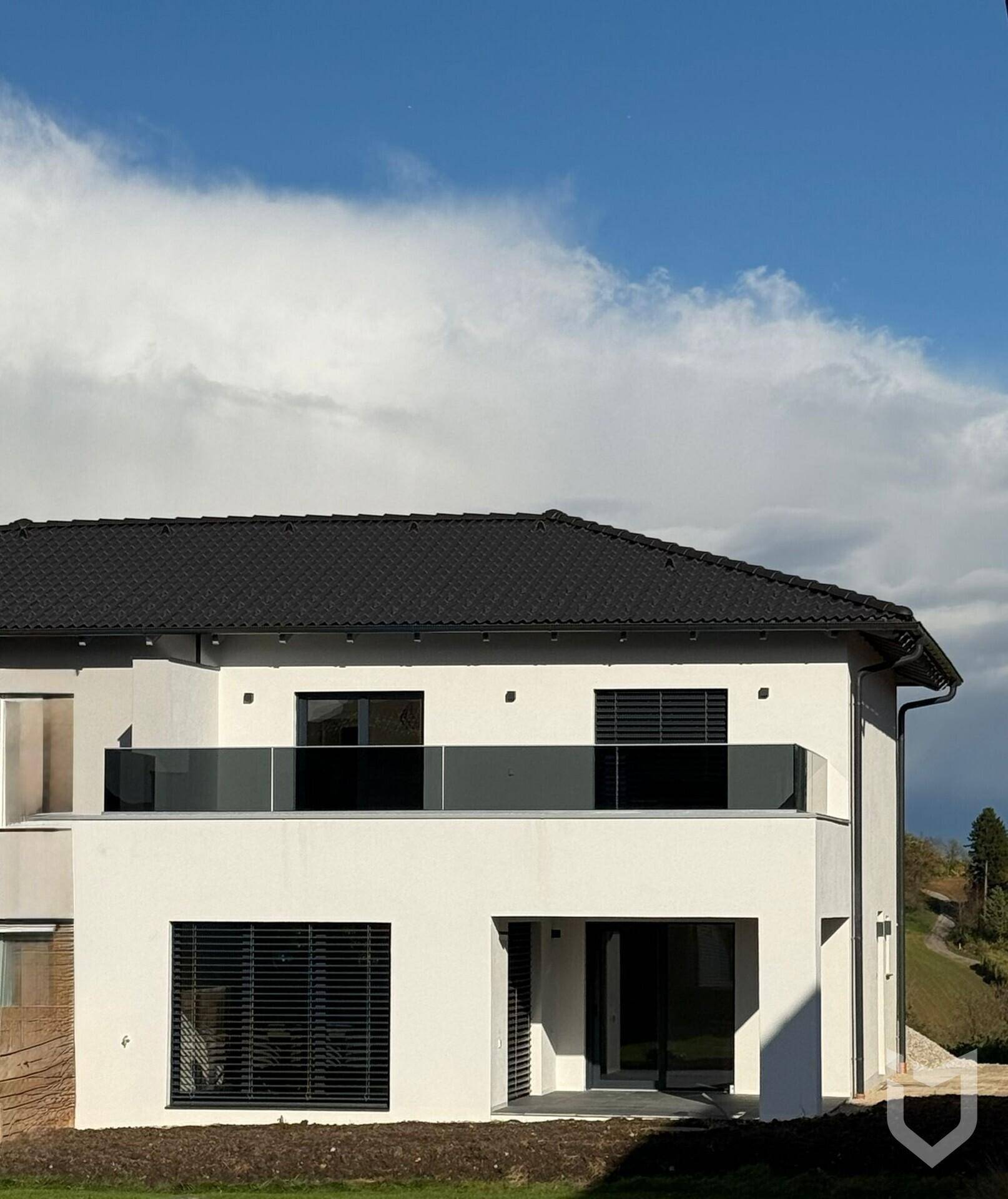 Moderne weiße Hausfassade mit schwarzem Dach und Balkon mit Glasgeländer.