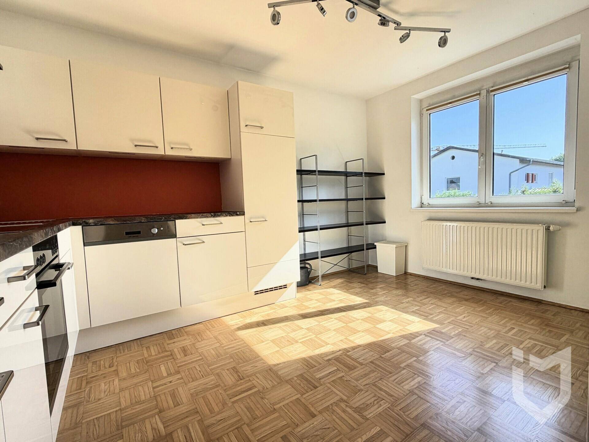 Küche mit hellen Schränken, Parkettboden und einem Fenster, das Tageslicht hereinlässt.