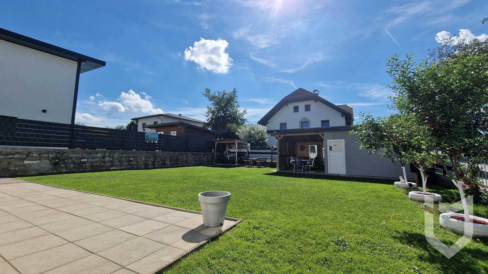 Weitläufiger Garten mit Rasenfläche, gepflasterter Terrasse und einem Gartenhaus.