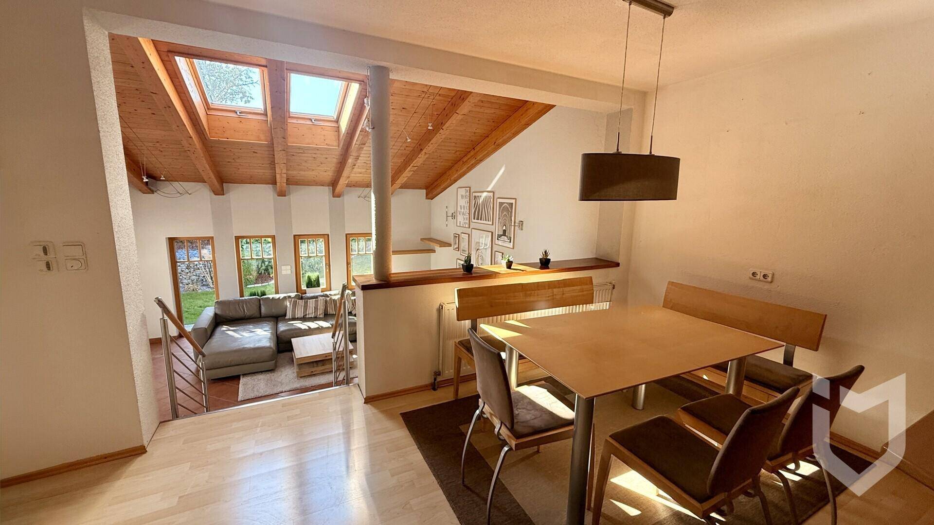 Heller Essbereich mit Holztisch und Blick auf das darunterliegende Wohnzimmer mit Holzdach und Dachfenstern.