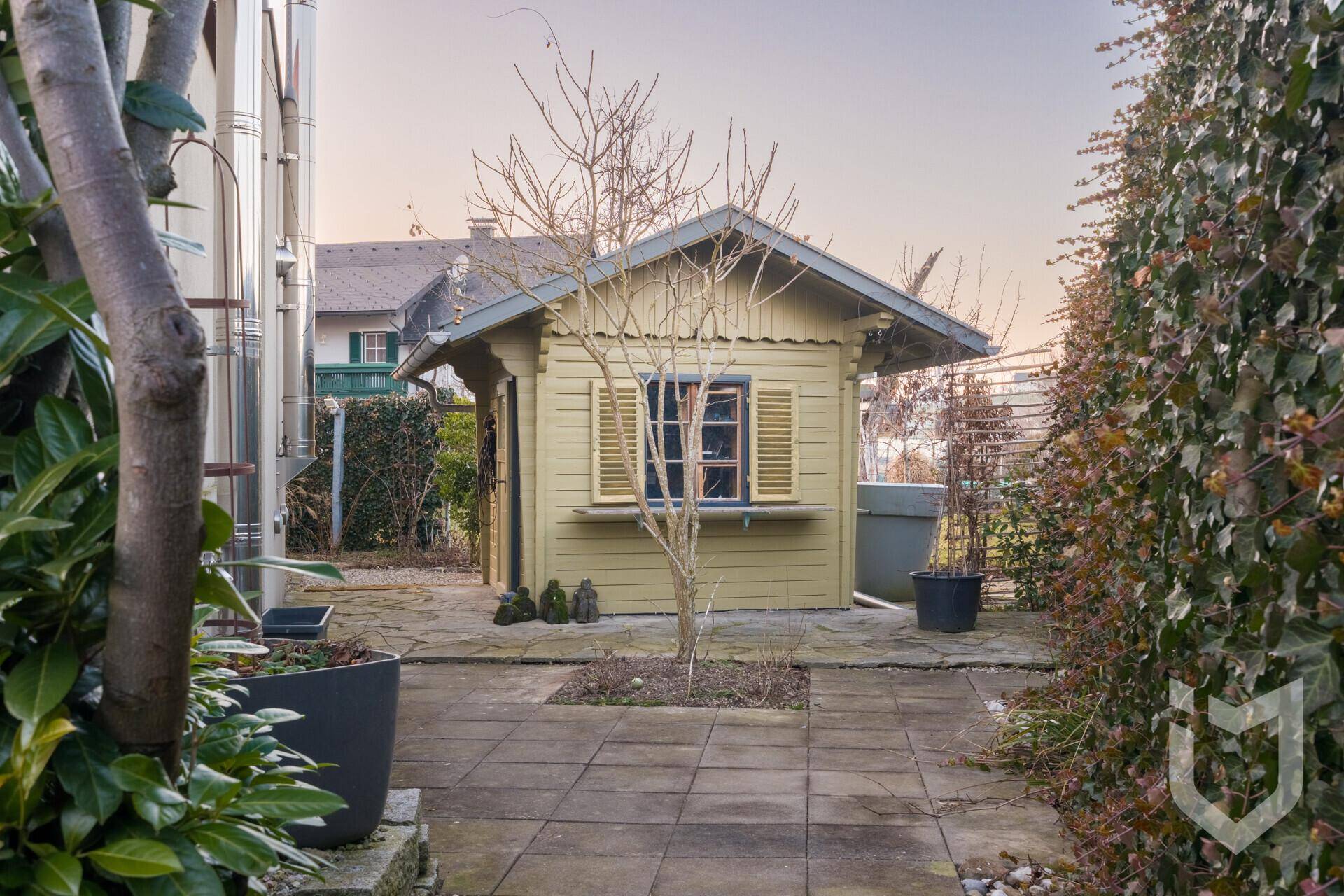 Gepflasterter Gartenbereich mit Gartenhaus, Bäumen und Pflanzen.