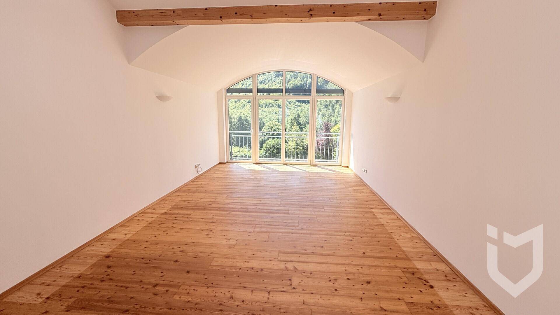 Lichtdurchfluteter Raum mit Holzdielenboden und einer großen Bogenfensterfront mit französischem Balkon.