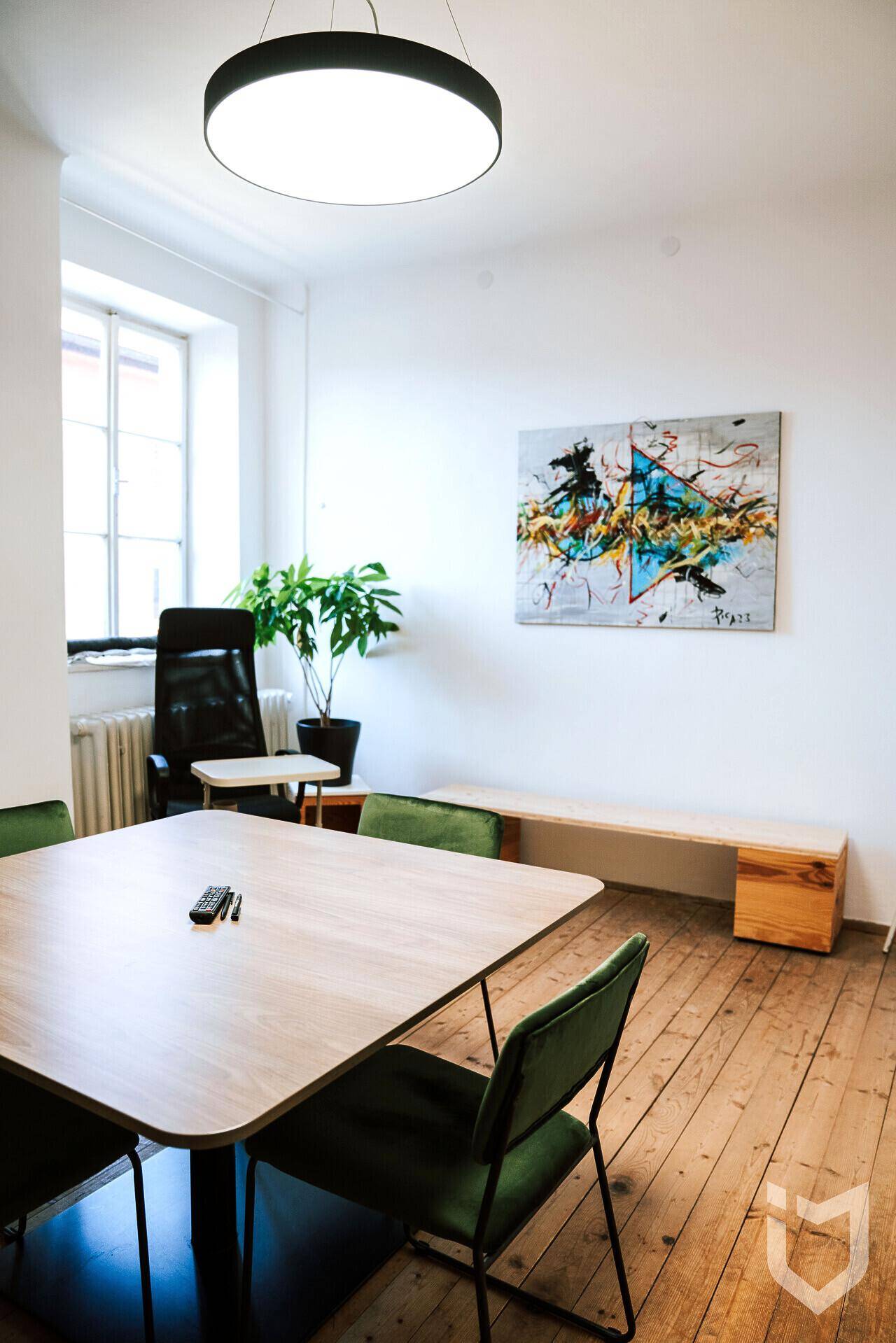 Besprechungsraum mit Holztisch, grünen Stühlen, Kunst an der Wand und einem großen Fenster für Tageslicht.