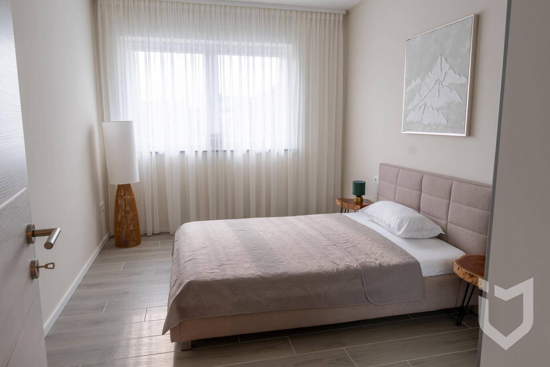 Helles Schlafzimmer mit Einzelbett, Nachttisch und großem Fenster mit Vorhängen.