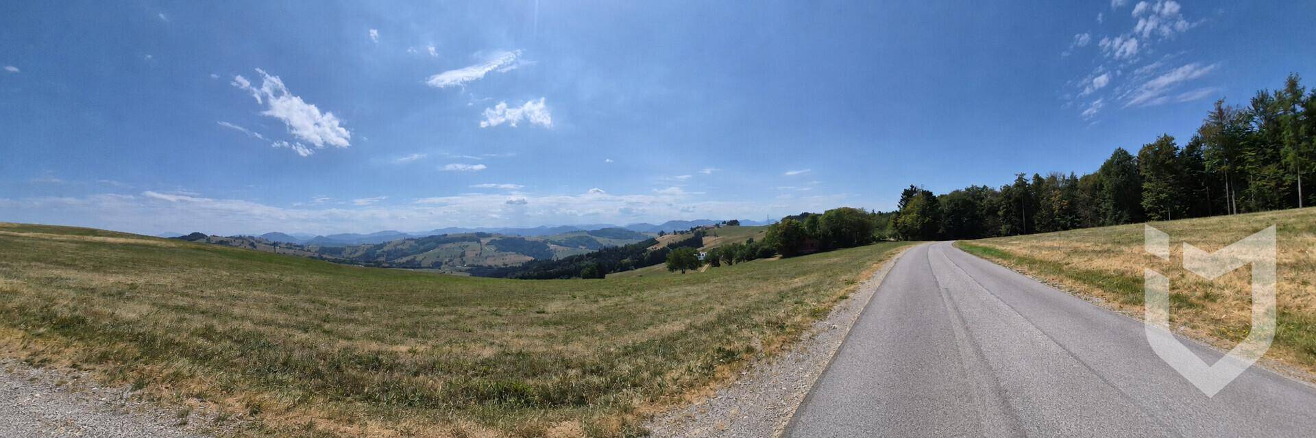 Panoramablick auf eine Landstraße, die sich durch eine weite grüne Landschaft mit Bergen am Horizont schlängelt.