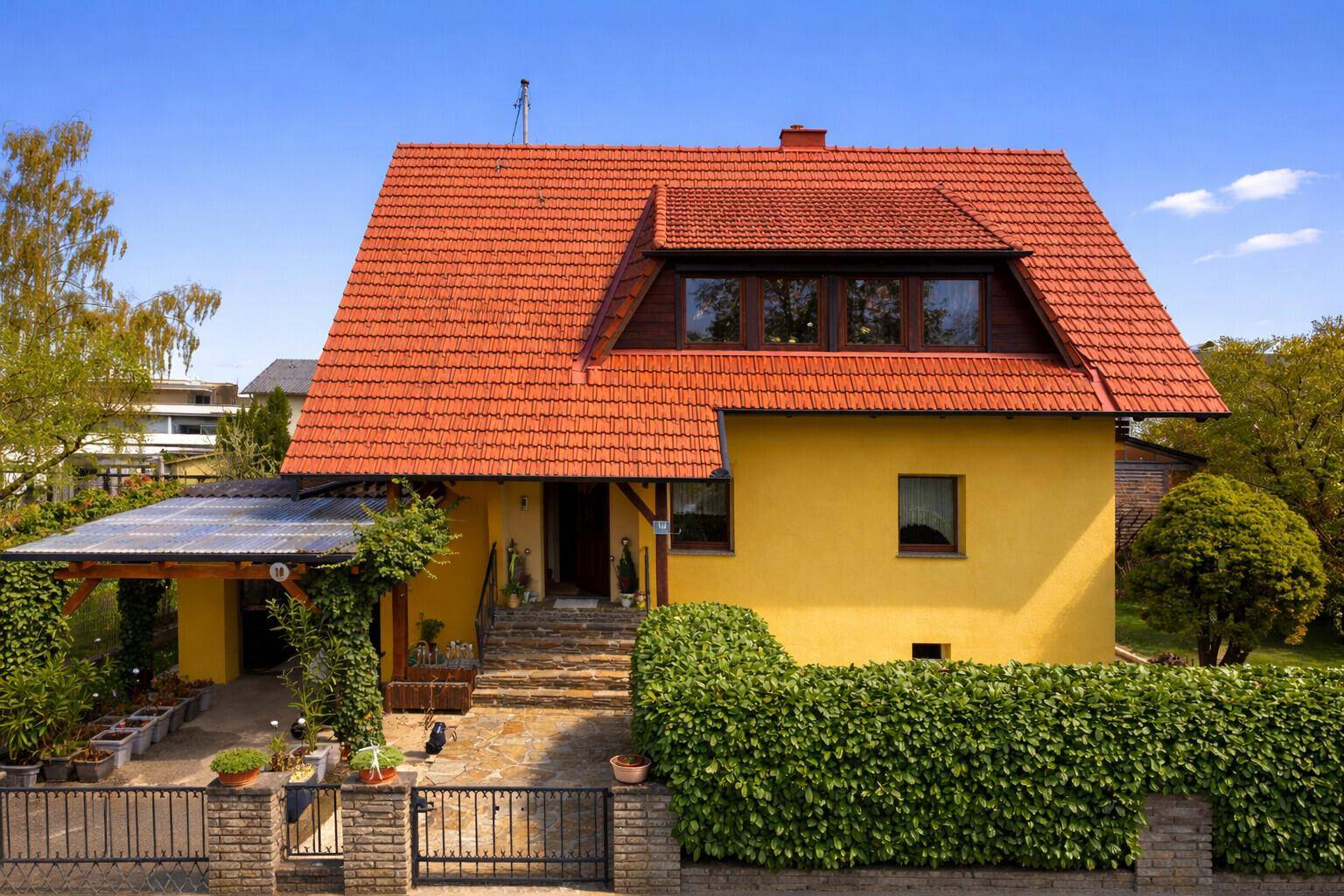 Vorderansicht des gelben Hauses mit rotem Dach, Carport und einem gepflegten Vorgarten mit Hecke.