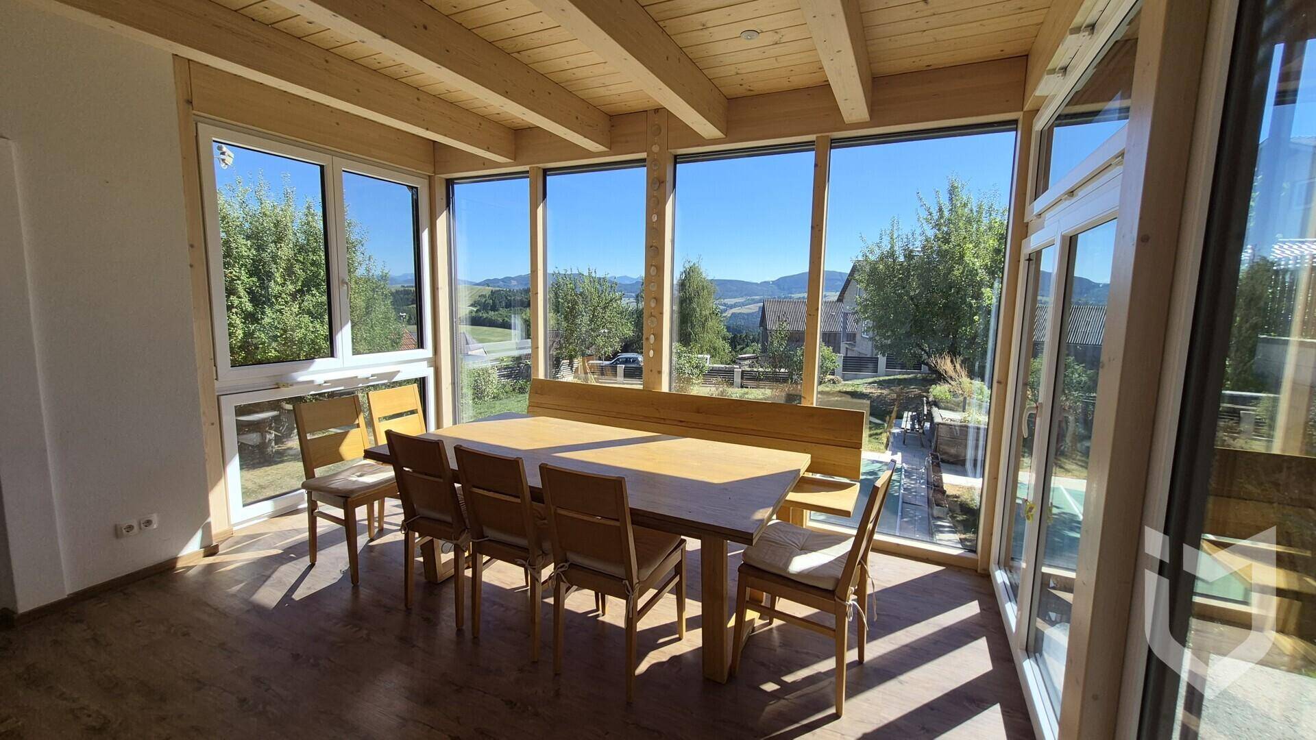 Heller Wintergarten mit Holzbalkendecke, großflächigen Fenstern und Esstisch mit Bergblick.